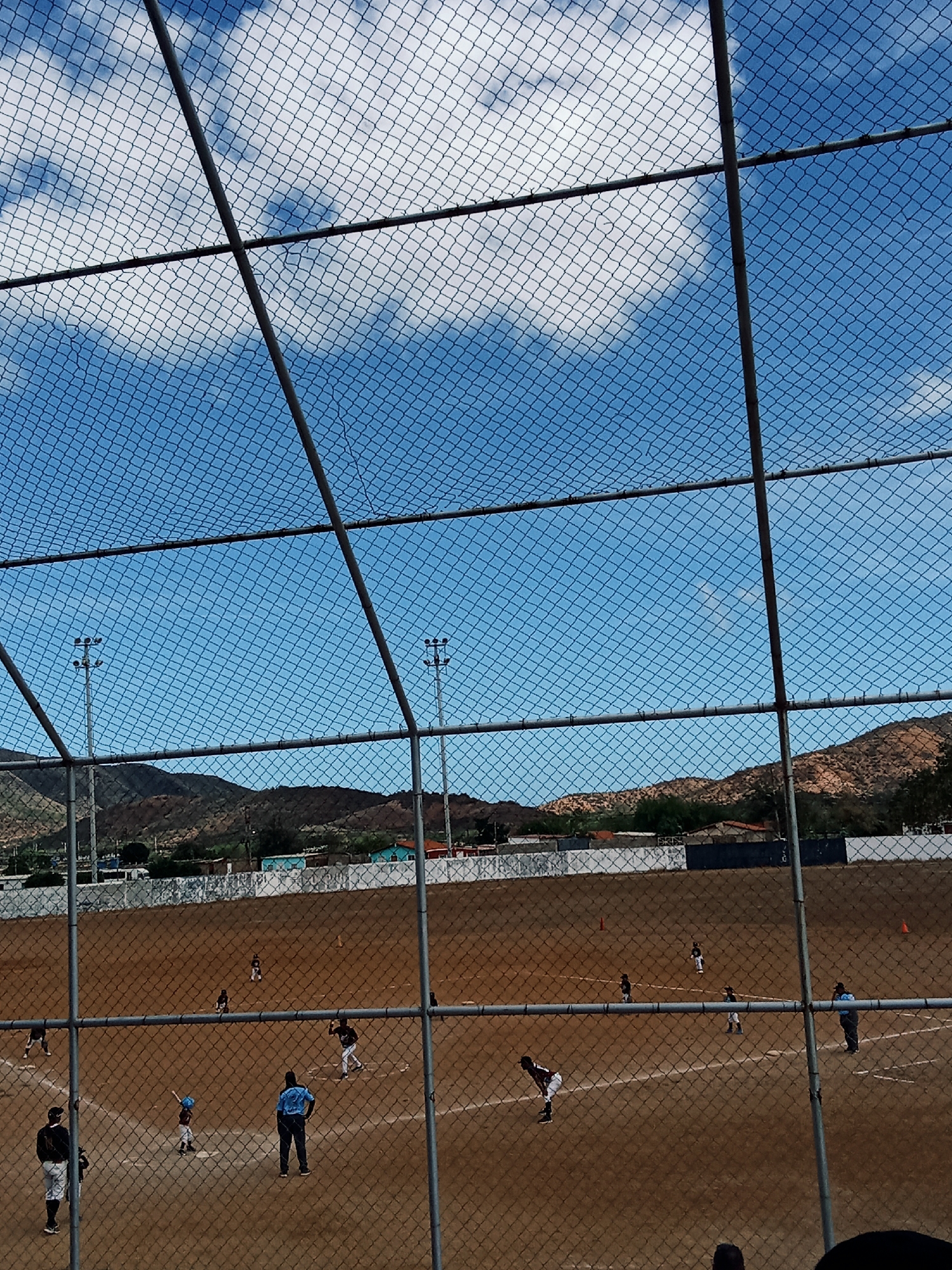 The image shows a baseball game in progress, viewed through a chain-link fence. A blue sky with scattered clouds is visible above the fence. Mountains are visible in the background beyond the field, along with some low buildings and a white wall. Several players are on the field, including a batter, a catcher, and other fielders. An umpire is standing near the batter. The game appears to be in a dirt field. The location is Juan Griego, Venezuela.
