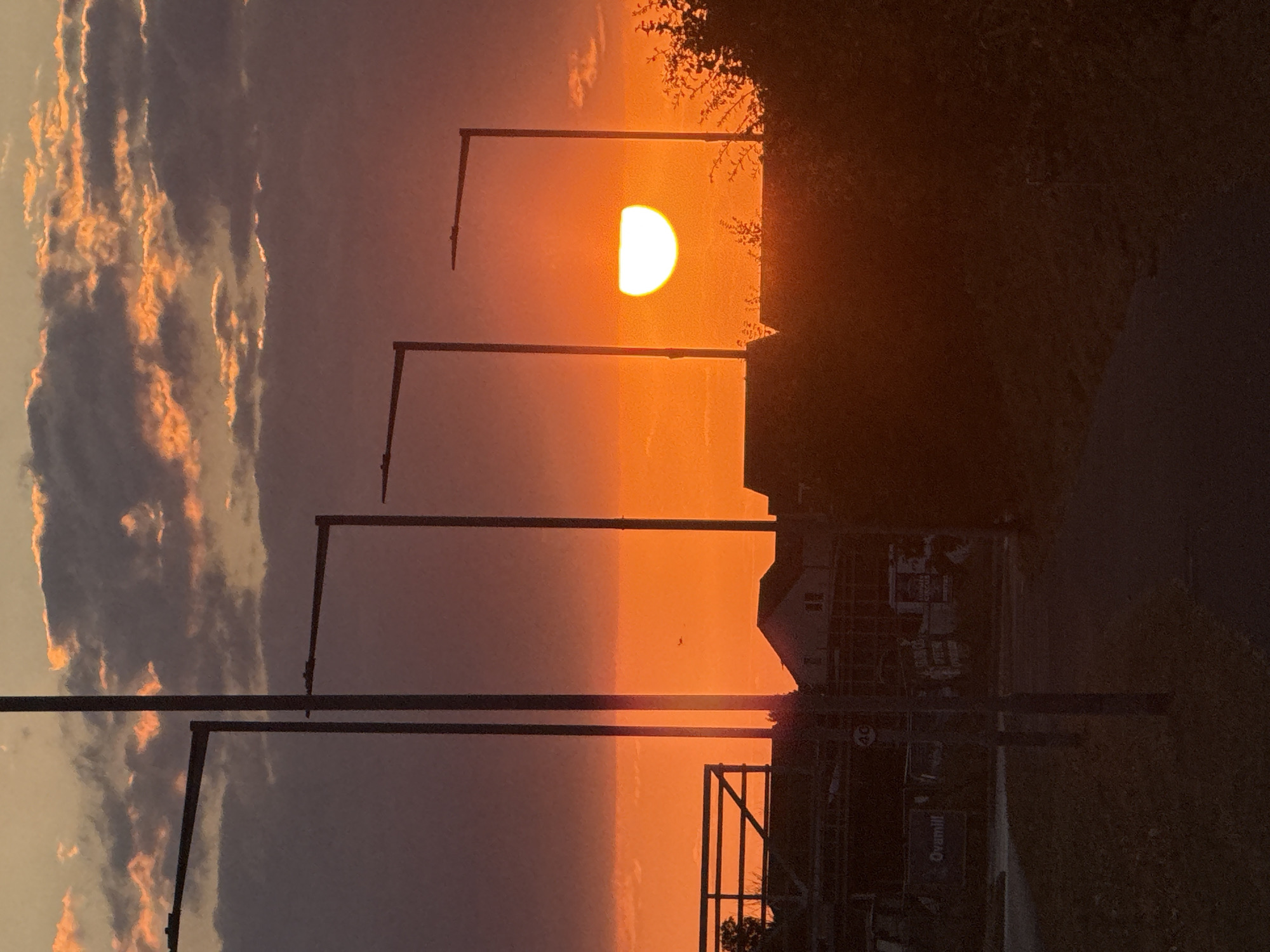 A silhouette of a rural or suburban landscape is presented against a vibrant sunset in Great Yarmouth, United Kingdom. The sky is filled with dramatic clouds, tinged with orange and purple hues as the sun, a bright, semi-circular orb, dips below the horizon. Several streetlights, tall and slender, stand in a line across the foreground, their dark forms stark against the colorful sky. In the middle ground, rooftops of houses and construction sites are visible, also silhouetted. A path or road leads into the distance, flanked by dark vegetation. The overall impression is one of twilight serenity.