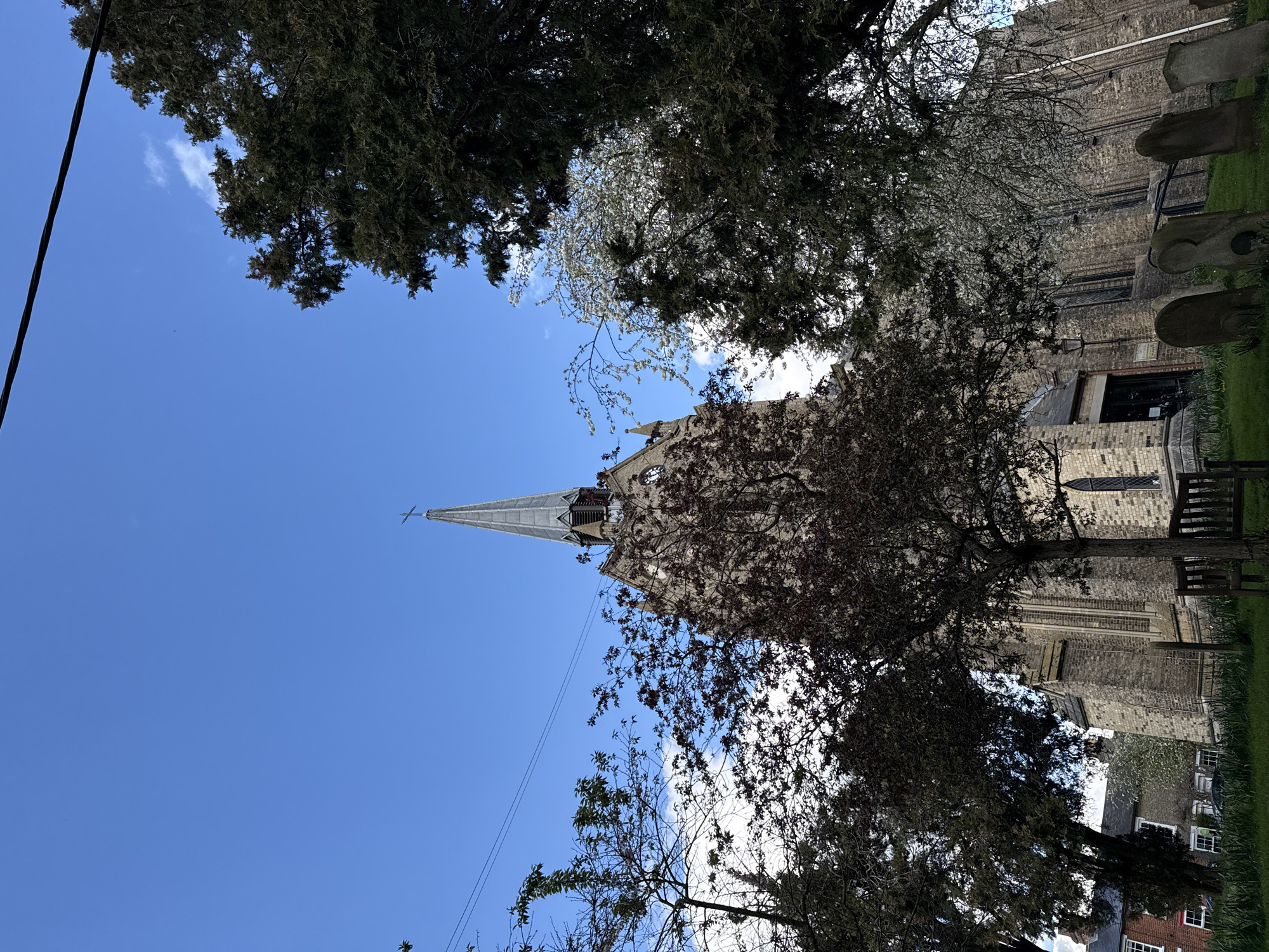 The image captures a view of St. Mary's Church in Bungay, East Suffolk, United Kingdom, framed by trees and a blue sky. The church's steeple rises prominently, with the main structure visible behind foliage. Tombstones are visible to the side of the building. Trees with a mix of green and brown foliage surround the building. A powerline can be seen. Grass and low buildings are visible.