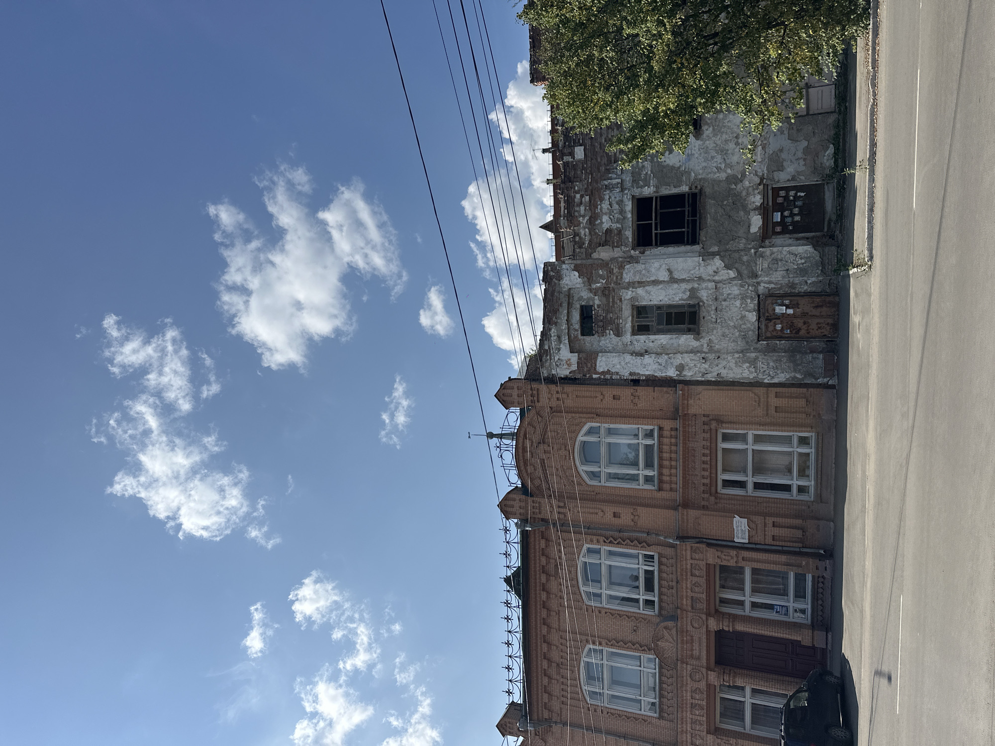 The image shows a street scene in Biysk, Russia, with two adjacent buildings. The building on the left is made of red brick and appears to be in good condition, with multiple windows and decorative architectural elements. The building on the right is in a state of disrepair, with crumbling plaster revealing brickwork and broken windows. A tree with green foliage stands to the right of the dilapidated building. Power lines are visible across the top of the frame, and a dark-colored car is parked to the left of the red brick building. The sky is blue with scattered white clouds.