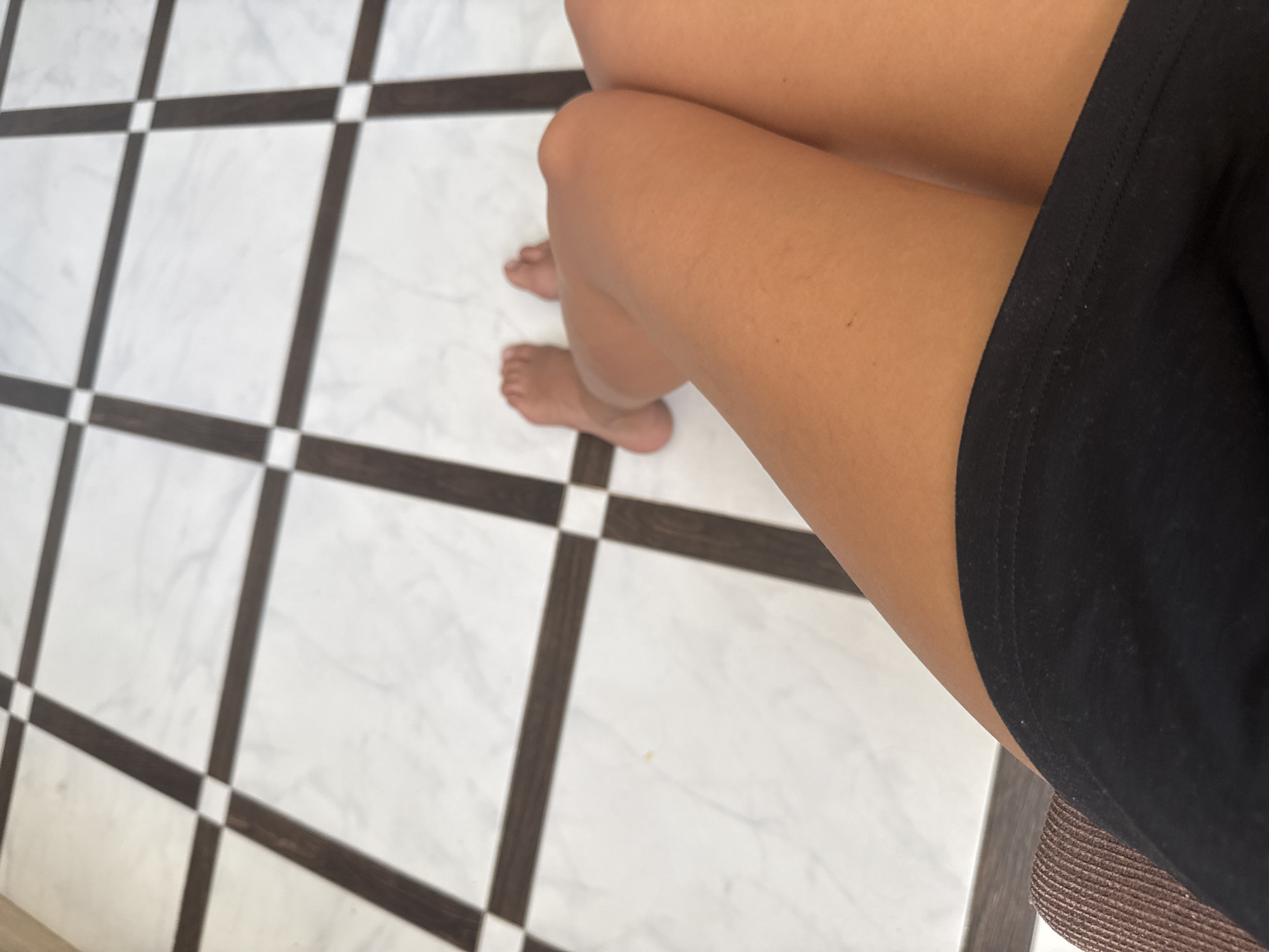 A person's legs are positioned in the foreground, with knees bent and feet visible in the background. The subject is wearing a black garment, likely shorts or a skirt, which is visible at the bottom of the frame. The background features a marble-tiled floor with a dark grid pattern. The scene appears to be indoors, potentially in a room with a decorative floor. The lighting suggests daylight, casting soft shadows. There is no visible indication of the location within the city of Chișinău, Moldova.