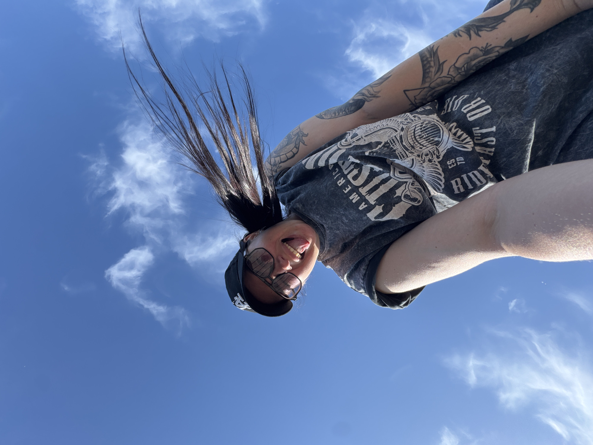 A low-angle photograph shows a woman with dark hair, wearing a black baseball cap and sunglasses, with arm tattoos, sticking her tongue out. Her hair is blowing upward against a blue sky with scattered clouds. She is wearing a gray t-shirt with a graphic design. The location is unspecified, in the city of Unknown, Unknown.