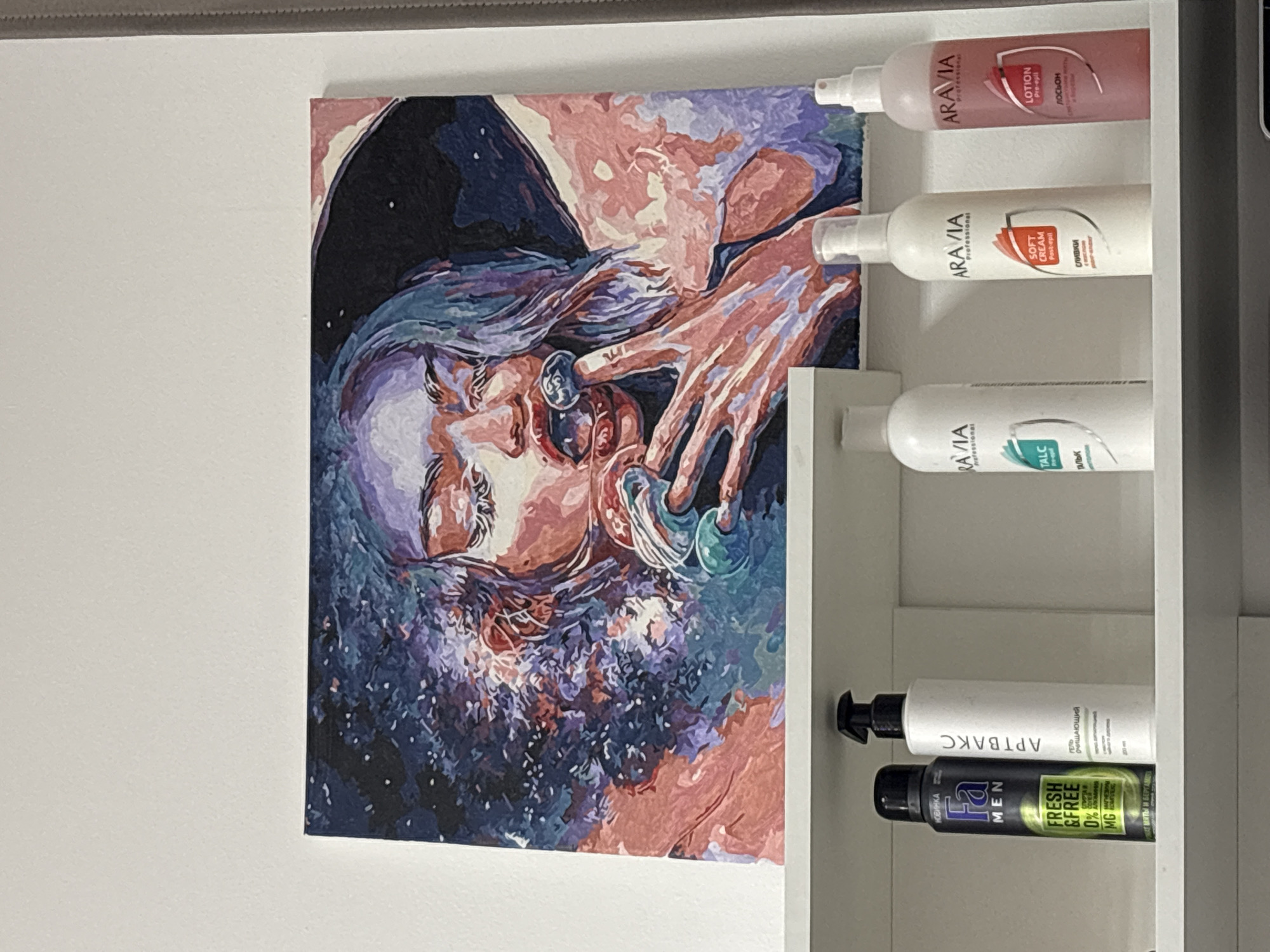The image displays an indoor scene, likely in Perm, Russia, featuring a vibrant painting alongside a shelf of personal care products. The main subject is a large, multi-colored painting depicting a woman's face and hands in an artistic, possibly paint-by-numbers style. Her eyes are closed, suggesting introspection or sleep, and her hair and surroundings are rendered in shades of purple, blue, and pink, with what appears to be a starry or abstract background. Her hands are near her face, possibly adorned with rings or other circular objects.
To the right of the painting, a white shelving unit holds several bottles of beauty and personal care products. On the top shelf, three "ARAVIA Professional" brand bottles are visible: a pink "LOTION Pre-epil," a white "SOFT CREAM Post-epil" (сливки), and another white bottle containing "TALC" (ТАЛЬК). The bottom shelf contains a white bottle labeled "АРТВАКС" (ARTWAX) "ГЕЛЬ ОЧИЩАЮЩИЙ" (CLEANSING GEL), and a green and black "Fa MEN" bottle, marked "НОВИНКА" (NEW) and "FRESH & FREE 0% MG." The Cyrillic text on the products reinforces the Russian location context. The painting is mounted on a plain white wall, illuminated by bright, even indoor lighting. No direct activity is shown beyond the display of art and products.