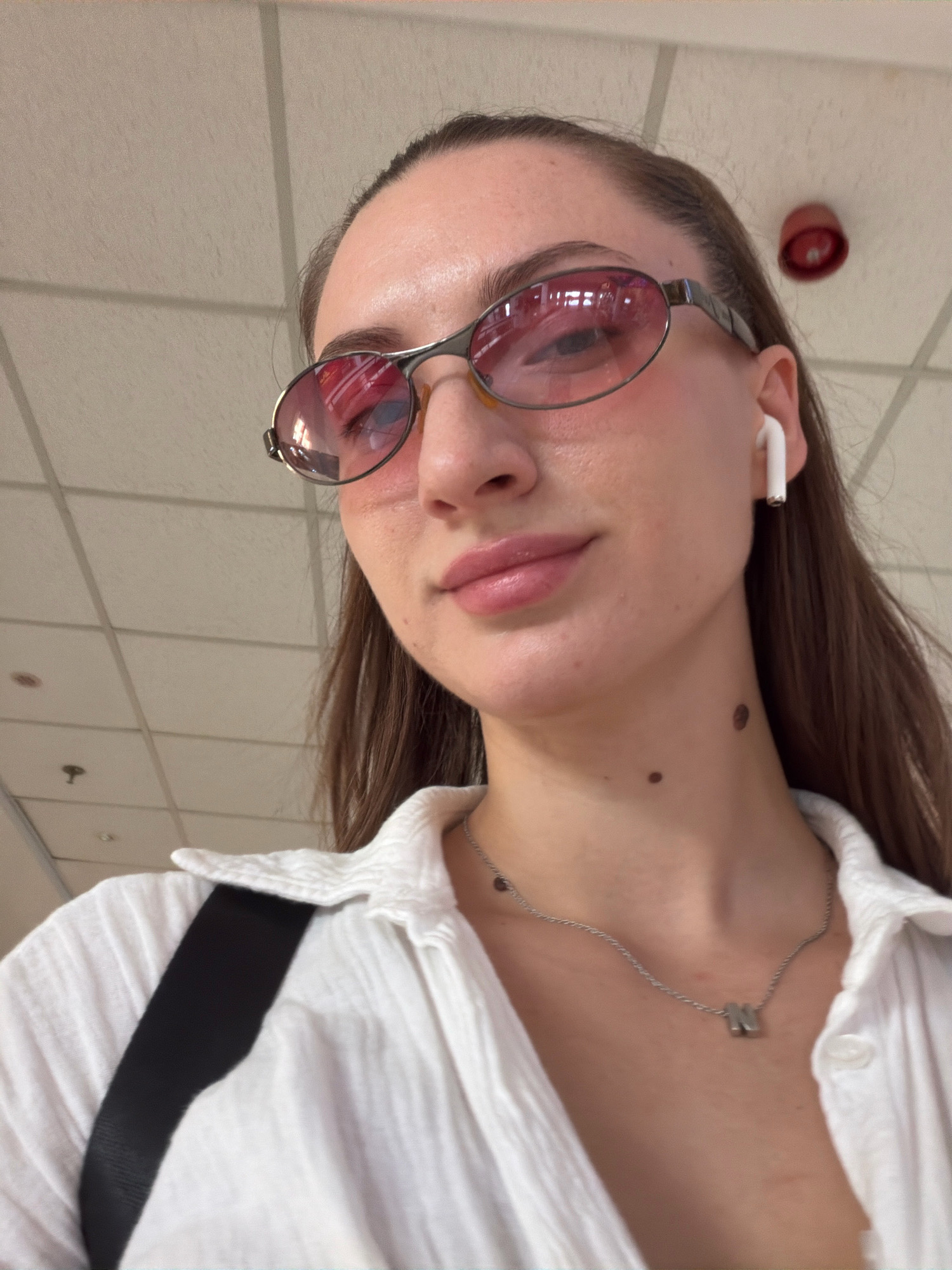 A close-up, selfie-style photograph captures a young woman from the chest up. She is wearing a white button-down shirt, a delicate silver necklace with a metallic letter 'N' pendant, and oval-shaped sunglasses with pink lenses. A white wireless earbud is visible in her right ear. The background appears to be an indoor ceiling with fluorescent lighting panels. There is a red circular object, possibly a fire alarm, mounted on the ceiling in the upper right portion of the image. The woman has fair skin and dark brown hair. There are two dark moles visible on her neck, one larger and darker than the other. The overall lighting suggests an interior space, and the ceiling structure indicates it could be a commercial or public building, such as an airport terminal or a shopping mall. The exact location within Sharm El Sheikh, Egypt, cannot be determined from the visual information present in the image.