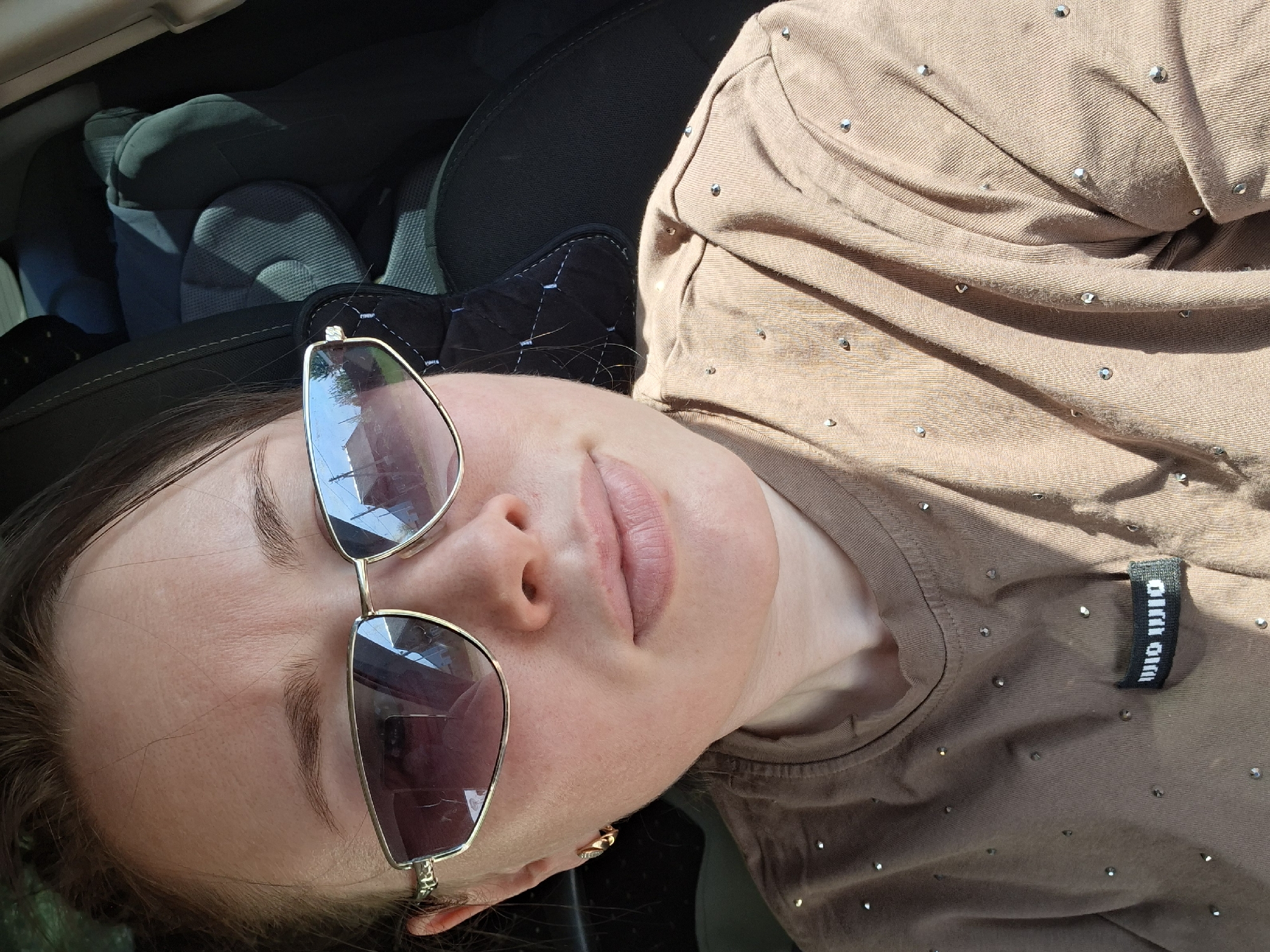 A woman is reclining in what appears to be a car seat. She is wearing sunglasses with gold frames. Her shirt is light brown with small, reflective accents. Part of the car's interior is visible in the background, including the car seat. The location is Unknown, Unknown.