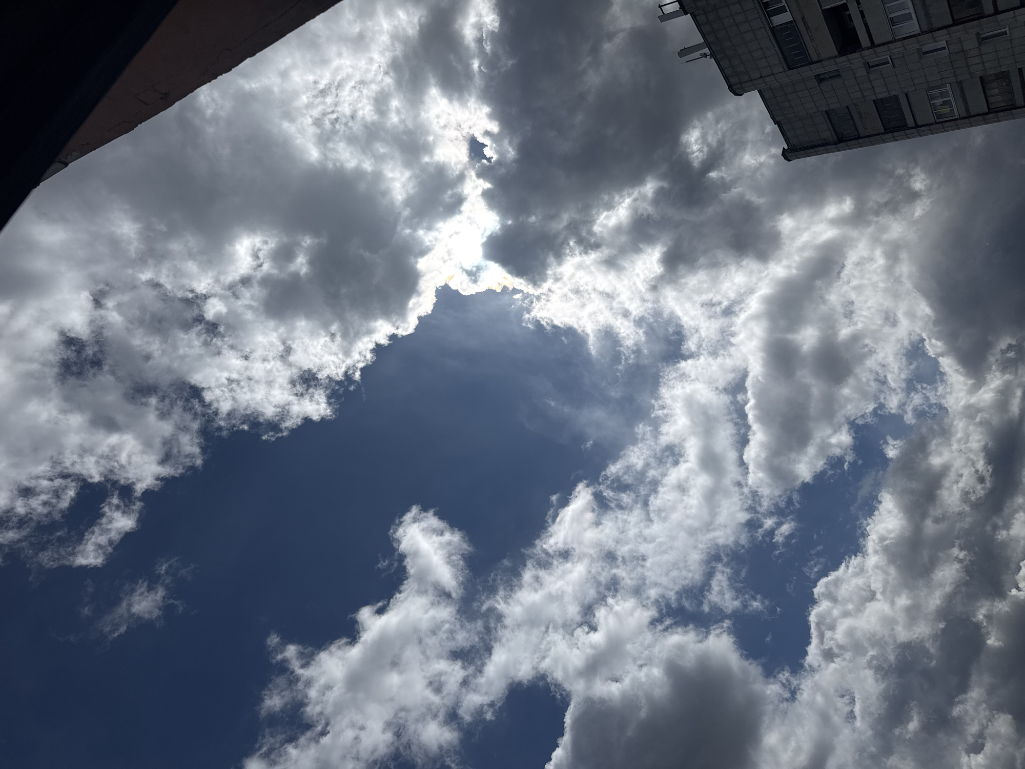 The image provides an upward-looking perspective of a partially cloudy sky, framed by elements of two buildings, set in Chaikovsky, Russia.
The scene is dominated by a dynamic arrangement of clouds and patches of deep blue sky. Large, billowing cumulus or stratocumulus clouds are prevalent, varying in density and illumination. In the upper-center, the sun is positioned directly behind a thick cloud mass, creating a brilliant, intensely bright halo that backlights the edges of the surrounding clouds, making them glow with a stark white light. Other clouds appear darker and grey, particularly on their undersides, indicating shadows. Several clear areas of deep blue sky are visible, especially towards the bottom and lower-right of the frame, interspersed with smaller, scattered white clouds.
On the upper-left, a dark, angular structure, possibly the shadowed roofline or eaves of a building, cuts diagonally into the frame. On the upper-right, a segment of a light-colored, multi-story building is visible. This building features a textured or tiled facade, with several dark, rectangular windows and what appear to be projecting ledges or fixtures. The architectural style suggests a functional, possibly Soviet-era design, consistent with its location in Russia.
The time of day is daytime, likely midday or early afternoon, given the high position and intensity of the sun behind the clouds. The weather is partially cloudy but sunny, creating strong contrasts between bright and shadowed areas. There is no specific activity or event depicted, nor is any visible text present. The image captures an everyday urban sky view.