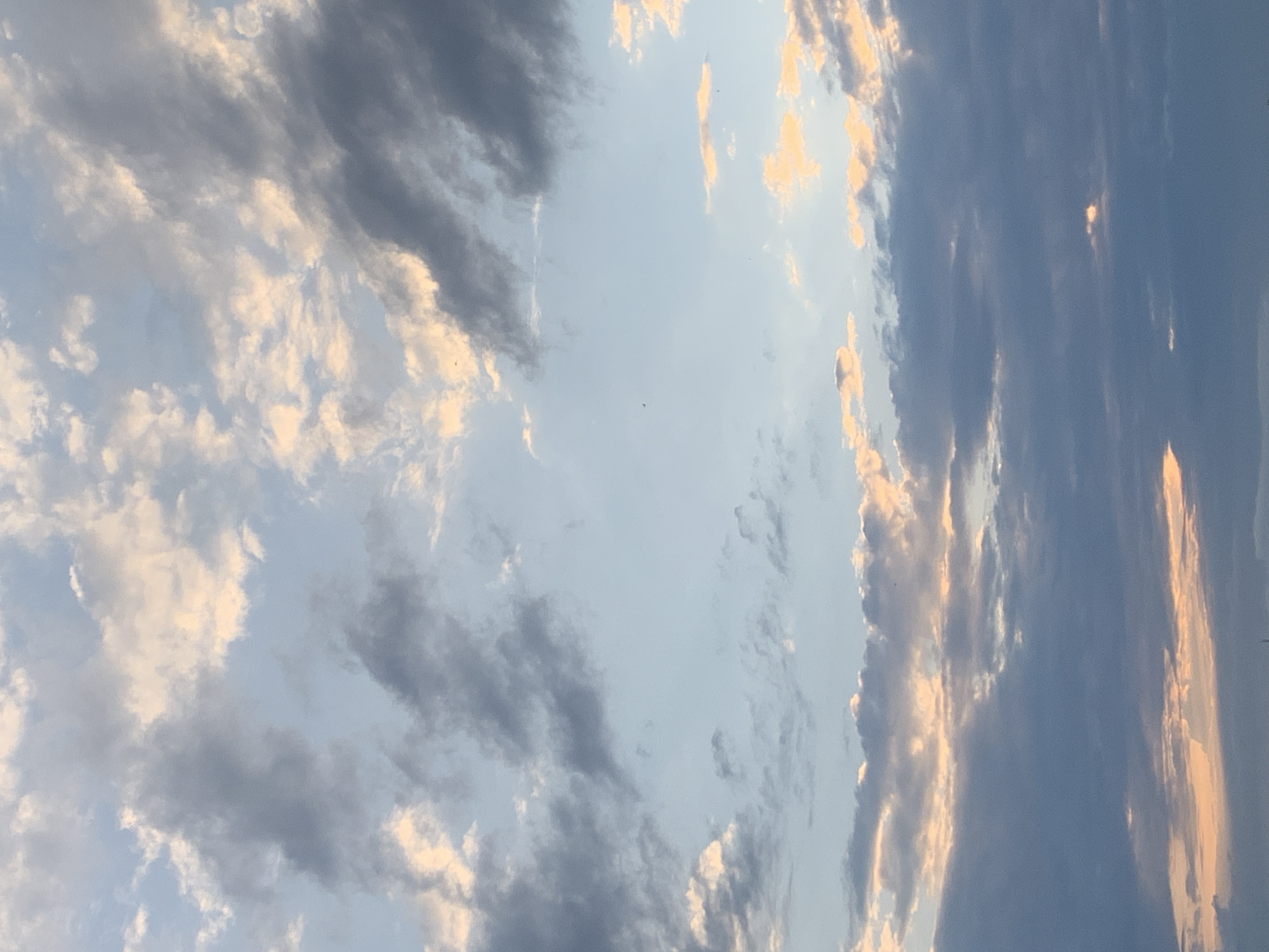 The image displays a sky filled with clouds. The clouds are a mix of dark gray and lighter white, with some areas tinged with a warm, peachy orange, suggesting either sunset or sunrise. The sky itself is a pale blue. There are no visible people, buildings, or specific landmarks that would indicate the location in Kolomyia, Ukraine. The image focuses solely on the atmospheric conditions of the sky.