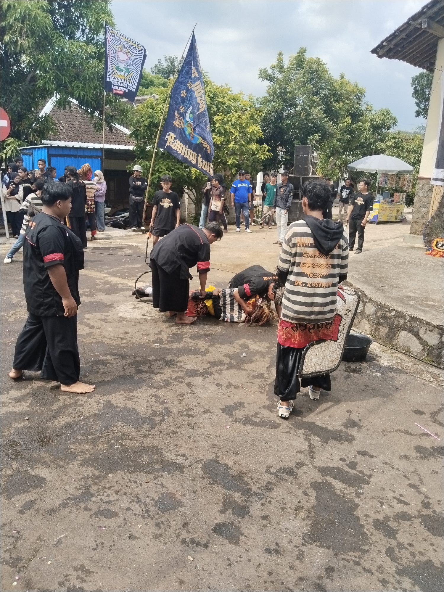 The image depicts a gathering of people in the city of Pendem, Indonesia. In the center of the frame, two individuals are attending to another person lying on the ground. All three are dressed in black and red. Two people stand to the left of the individuals on the ground, and one holds a Sembrani shield. Several other individuals are standing in the background, watching the scene. Two flags are raised on flagpoles, and several buildings and trees are visible. The ground is paved and has a mottled appearance.