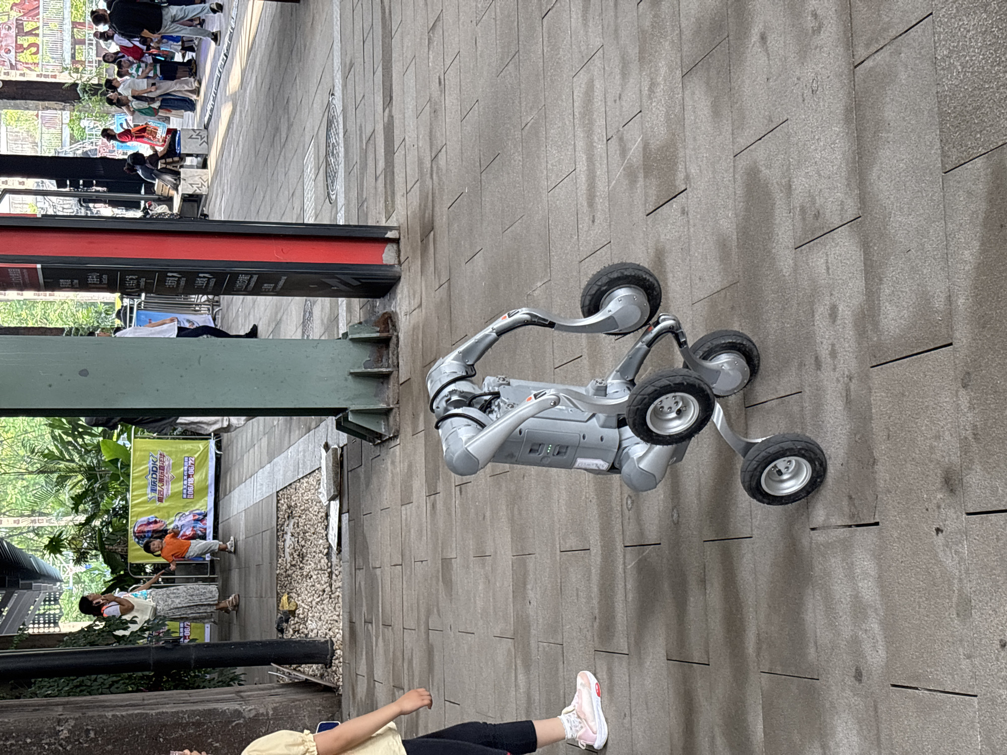 The image depicts a robot with multiple wheels, appearing to be a wheeled robot designed for locomotion, standing on a paved outdoor walkway. The robot is gray and silver, with articulated arms and a cylindrical body. In the background, there are several people walking and standing, indicating a public area. There is also a green banner with Chinese text, suggesting a commercial or event space. The walkway is made of large, rectangular stone tiles, some of which appear wet. The overall environment suggests a bustling urban setting. The specific location in Dadukou, China, is not explicitly indicated by any visible landmarks or signs in the image, but the presence of Chinese text on the banner implies a Chinese-speaking region.