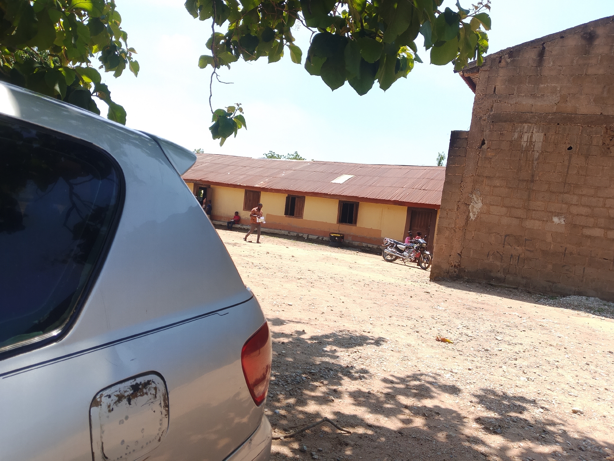 This image depicts an outdoor scene in Madalla, Nigeria, under bright daylight with a clear blue sky. In the immediate foreground, the rear quarter of a silver vehicle, likely an SUV or minivan, is partially visible on the left. It features a dark-tinted rear window, a visible fuel cap area on the fender with significant rust and peeling paint, and a thin dark pinstripe running along its side. Overhead, green tree branches with dense foliage extend into the frame from the top, casting dappled shadows across the sandy-dirt ground.
The midground is dominated by the uneven, light brown dirt ground, strewn with small stones and pebbles. A male individual, wearing a patterned shirt and light trousers, walks from left to right across this ground, holding an item in his left hand. To his left, near a building, a smaller figure in a red shirt is seated. Further right, a yellow portable generator rests on the ground near a building wall. A dark-colored motorcycle is parked on the far right, adjacent to a group of people, one of whom is wearing a pink top.
In the background, a long, single-story building stretches across the scene. It features light yellow or cream-colored walls with a darker base and multiple rectangular windows fitted with wooden shutters. The building is topped with a weathered, reddish-brown corrugated metal roof. To the far right, a tall, unfinished structure made of rough, brown concrete blocks rises, showing signs of wear and incomplete construction.
