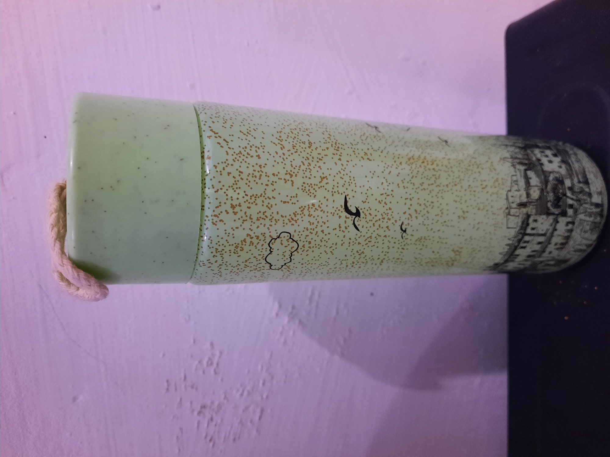 A light green cylindrical mug, decorated with a speckled pattern and illustrations of birds, clouds, and a cityscape, stands on a dark surface against a textured, purple-hued wall. A light-colored rope loop is attached to the top of the mug. The mug is approximately three-quarters of the way into the frame from the right side. The wall behind it is uneven and shows areas of discoloration. The dark surface appears to be a shelf or tabletop. The image captures a still life of the mug. The location in the city of Lhokseumawe, Indonesia, is not discernible from the visual information provided in the image.