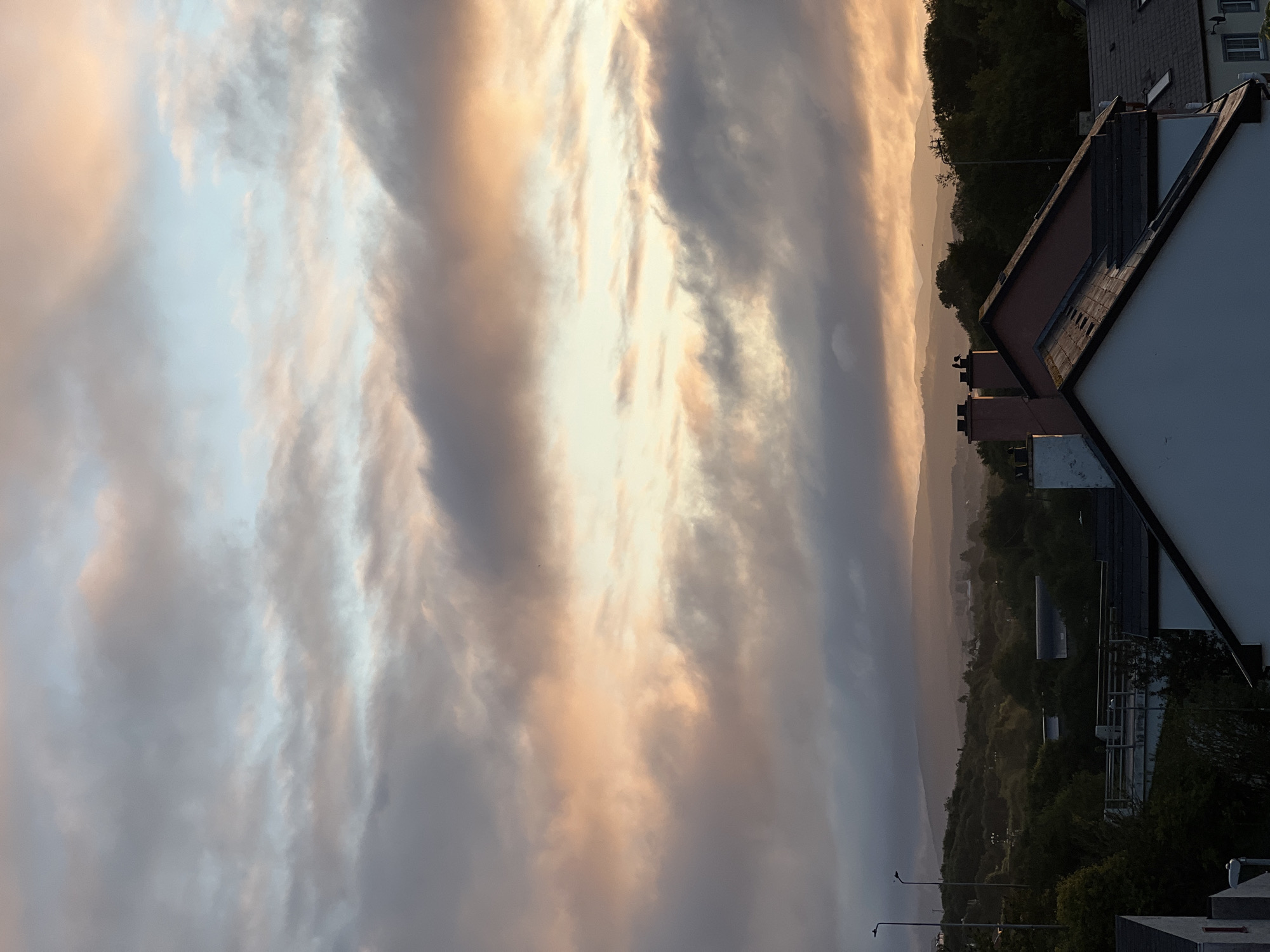 The image is a landscape view of Mountcharles, Ireland, at dusk. The sky is filled with dramatic, textured clouds in shades of pink, orange, gray, and blue, suggesting a sunset. In the foreground, the roofs of several houses are visible, with chimneys on some of them. Two birds are perched on one of the chimneys. Beyond the houses, a rolling, tree-covered landscape stretches into the distance, with hills or mountains silhouetted against the glowing sky. The overall atmosphere is serene and evocative of the natural beauty of the Irish countryside at twilight.