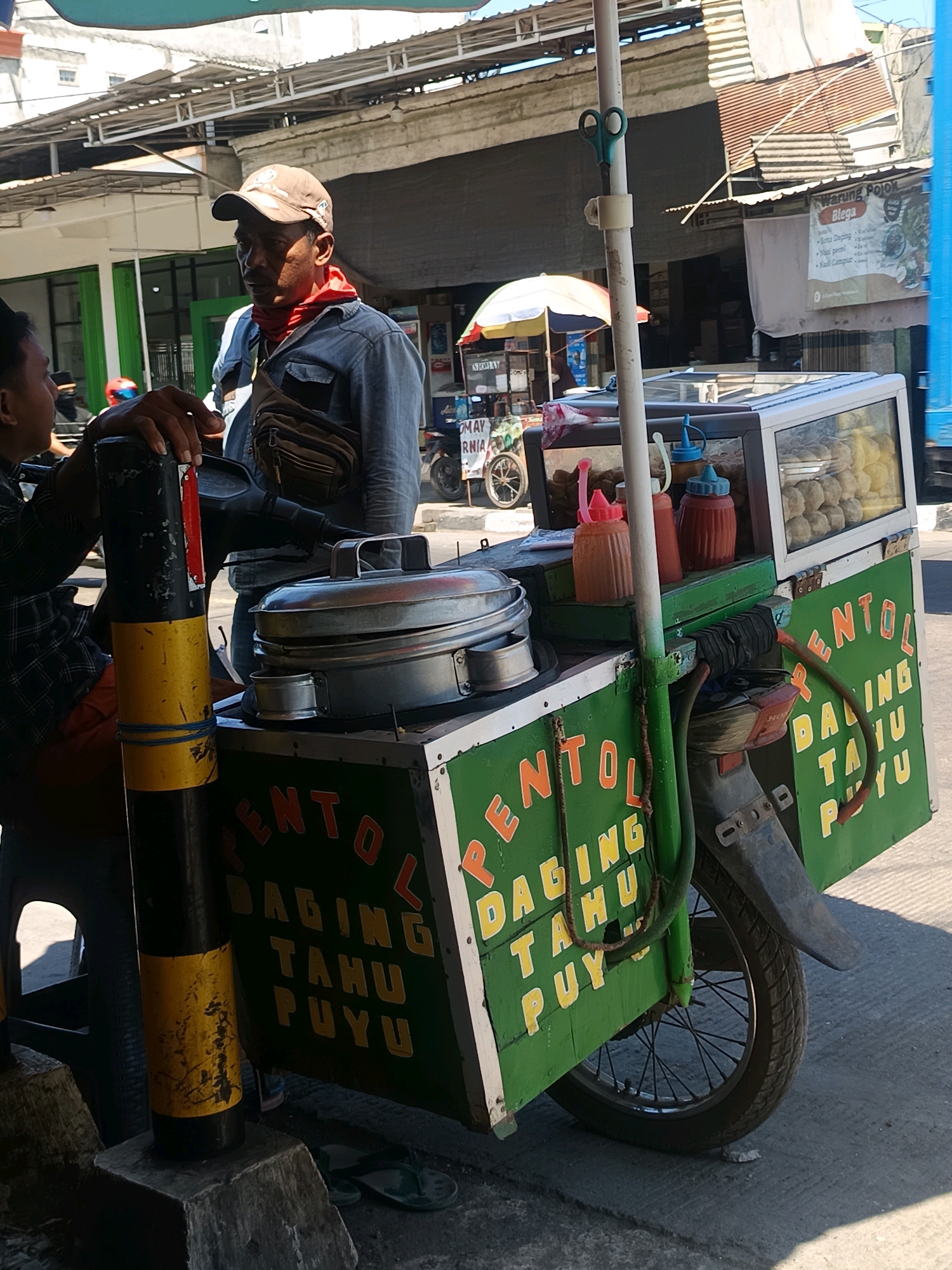 A street food vendor in Blega, Indonesia, operates a mobile cart selling "Pentol Daging Tahu Puy." The vendor, a middle-aged man wearing a blue denim shirt, a brown baseball cap, and a red bandana around his neck, stands near his cart. He has a fanny pack across his chest. The cart itself is green with text advertising the food in orange and yellow. It is equipped with a glass display case showcasing spherical food items, likely the "pentol," and several condiment bottles. A metal steamer sits on the cart. The cart is attached to a motorcycle, indicated by the visible wheel and fender. The scene takes place outdoors on a paved surface, with buildings and other street vendors visible in the background. Sunlight casts shadows, indicating it is daytime. A pair of dark flip-flops rests on the ground near the cart.