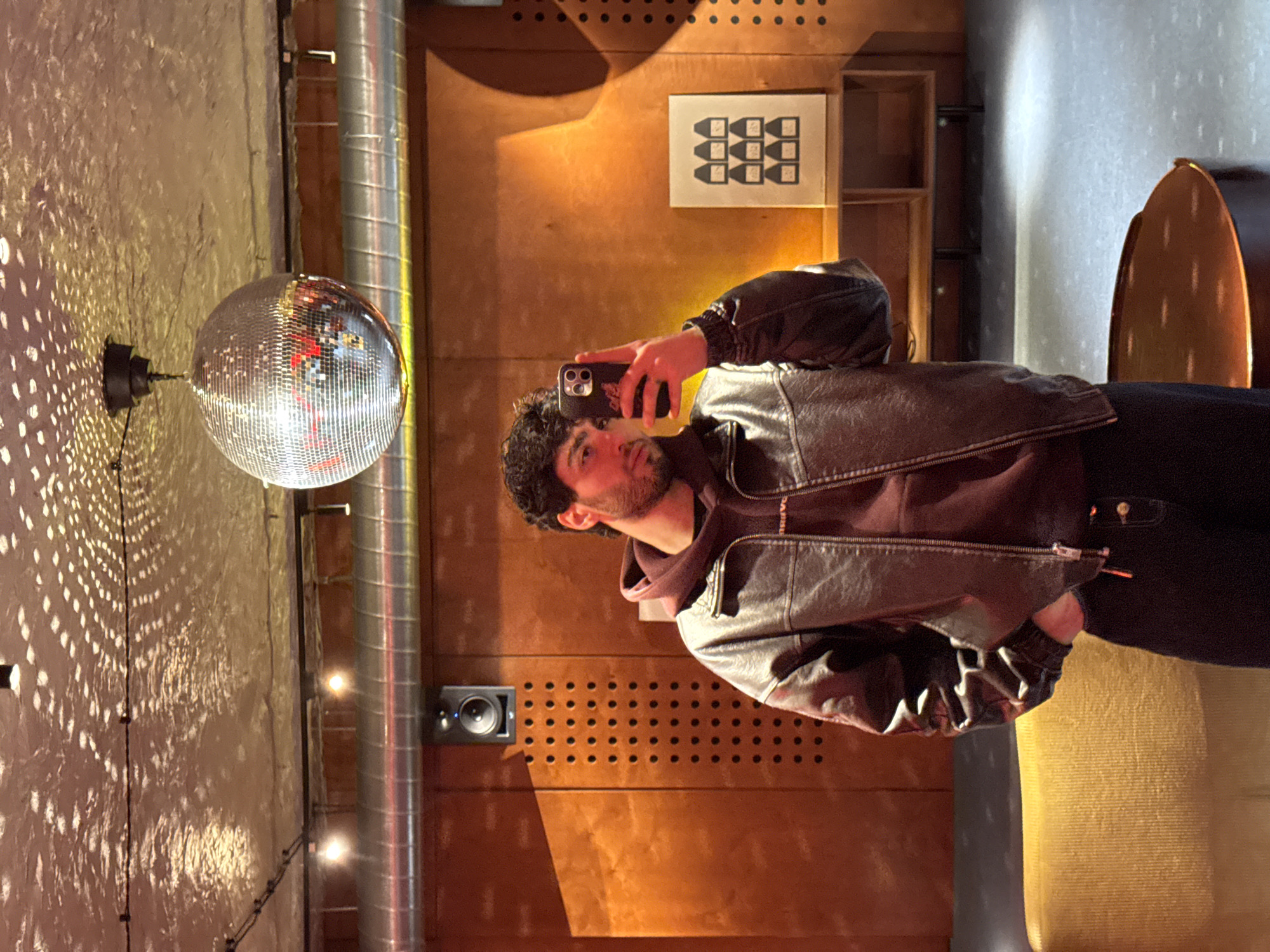 A man is taking a selfie in what appears to be a bar or club. He has curly dark hair, a beard, and is wearing a dark leather jacket over a hooded sweatshirt. He is holding a phone in his right hand to take the picture. In the background, there is a disco ball hanging from the ceiling, casting reflections onto the wall. A metal pipe runs vertically along the wall. There is wooden paneling on the wall behind the man with decorative holes and a small speaker. A framed piece of art with a geometric design is also visible. The location may be in Yerevan, Armenia.