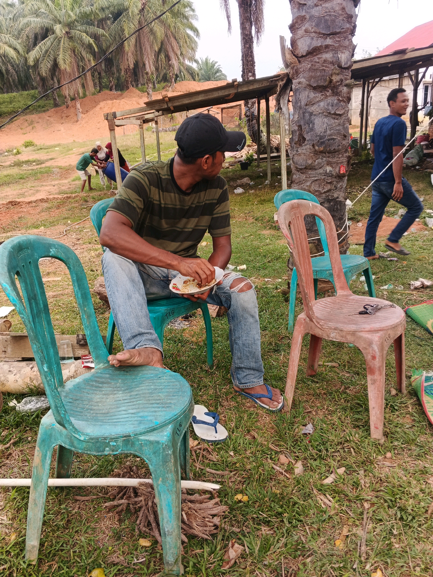 In Brangan Mulya, Indonesia, a man wearing a black cap and a striped t-shirt is seated on a teal plastic chair, eating from a plate in his left hand. He is wearing blue jeans with a ripped knee and blue flip-flops. His right leg is crossed over his left. In the background, a man in a blue t-shirt and jeans walks away, and further behind, a group of people are gathered around a large blue barrel. The scene is outdoors, with palm trees and a makeshift structure visible in the background. The ground is covered with grass and dirt, and several other plastic chairs are present.