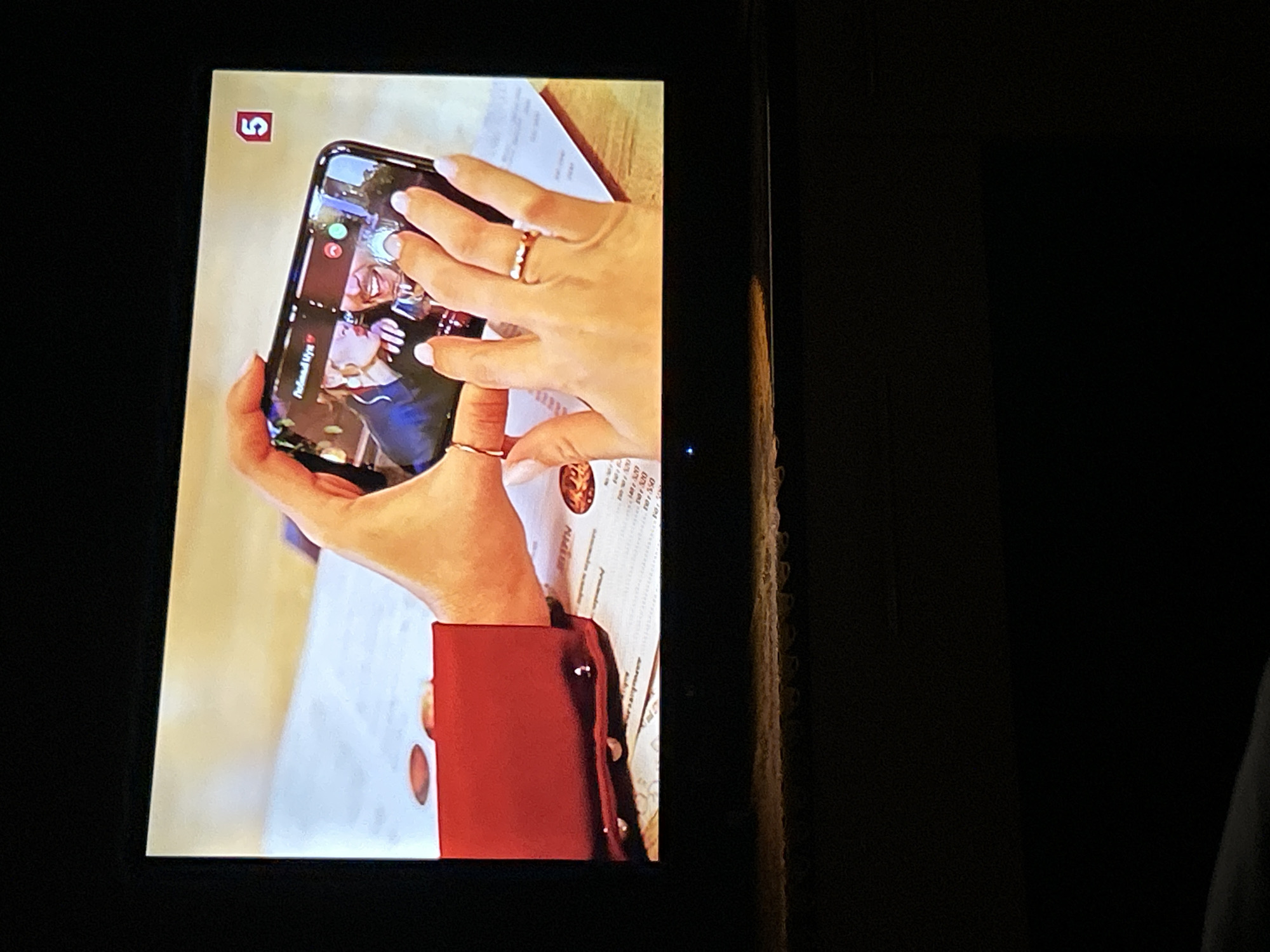 The image shows a close-up view of a television screen displaying a video call on a smartphone. Two hands are holding the smartphone, with the right hand's fingers visible pressing the screen. The video on the phone shows a man and a woman embracing, with the woman kissing the man's cheek. The woman in the video is wearing large hoop earrings. On the phone screen, text reads "Любимый Муж" which translates to "Beloved Husband." A red button with a white "5" logo is visible in the upper right corner of the television screen. The television screen is dark around the edges. The setting is indoors, with a blurred background suggesting a table with papers and a red fabric garment in the foreground. The overall scene suggests a personal moment being shared via video call, possibly with a wedding or romantic theme. There is no information in the image to indicate a location in Gelendzhik, Russia.
