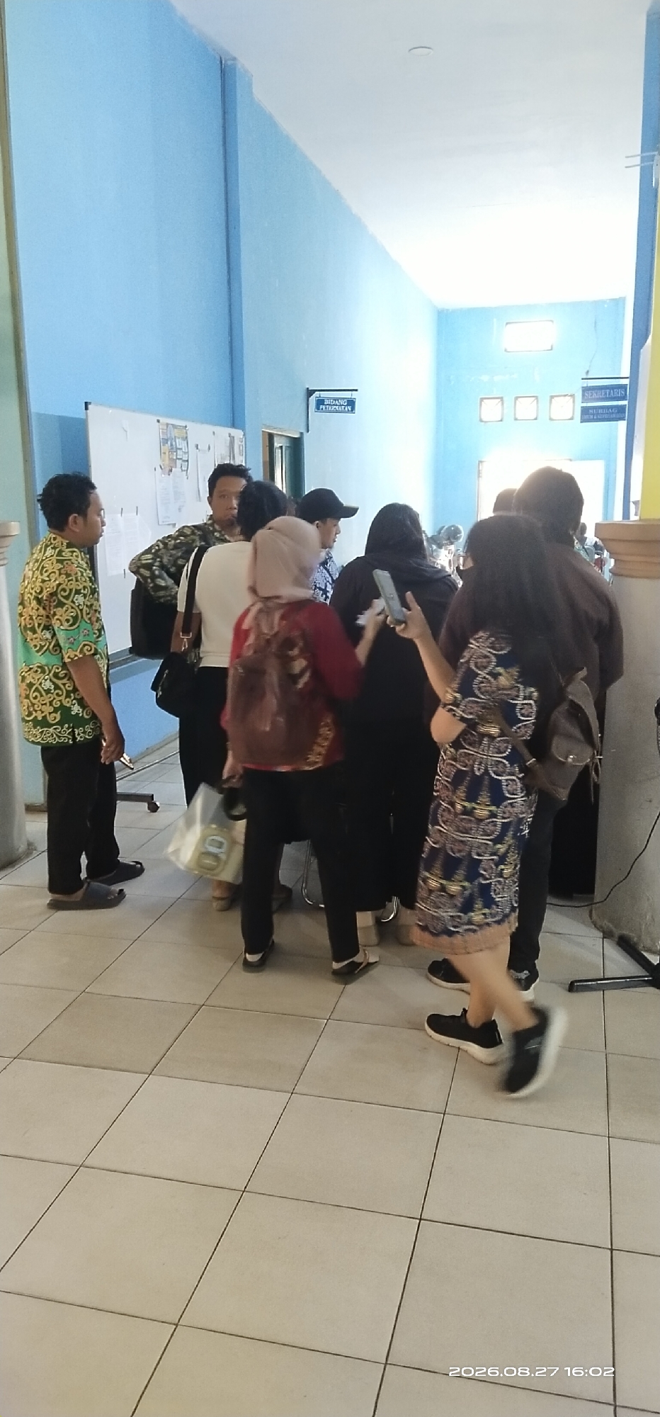 The image depicts a group of people standing in a hallway. On the left, a man in a green and gold patterned shirt and black pants stands facing left, looking away from the camera. Next to him, another man in a patterned shirt and black pants faces the camera, with a dark bag over his shoulder. Several other individuals are visible in the background, some with their backs to the camera, including a woman in a red headscarf and patterned backpack, and a woman in a blue patterned dress. Signs are visible on the light blue walls, one reading "BIDANG PETERNAKAN" and another "SEKRETARIS". The floor is tiled with a grid pattern of light-colored squares. The lighting suggests an indoor environment, likely during the day. The timestamp in the lower right corner indicates the date and time as 2026.08.27 16:02.