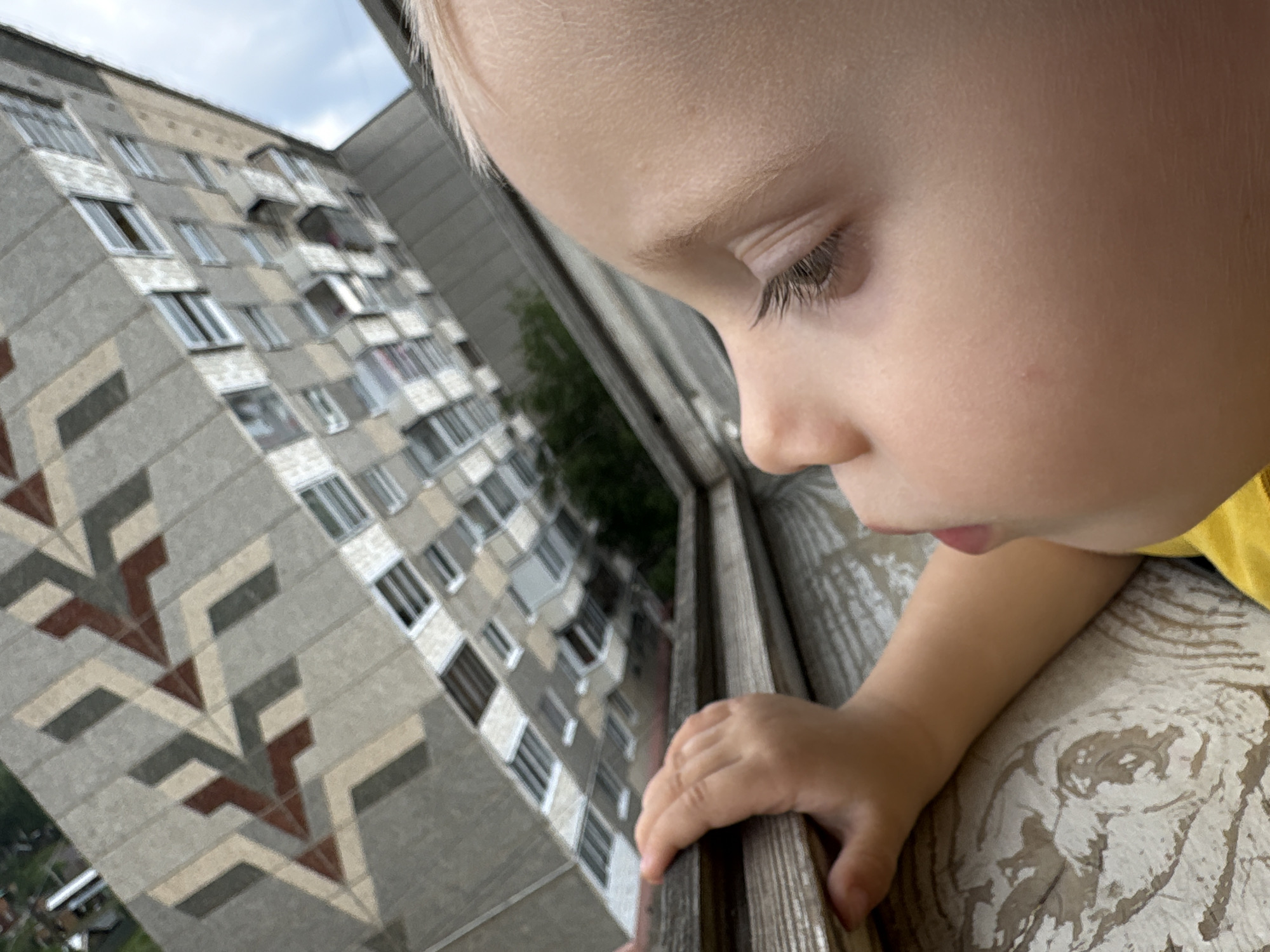 A child's face, framed by fair hair, is captured in a close-up profile view as they look out of a window. The child's right hand grips the wooden frame of the window. Reflected in the glass of a large, multi-story building across from the window, the child's face is partially visible. The building's facade features a geometric pattern in shades of gray, beige, and brown, with numerous windows and balconies. The scene appears to be from Chaikovsky, Russia, a city known for its Soviet-era architecture.
