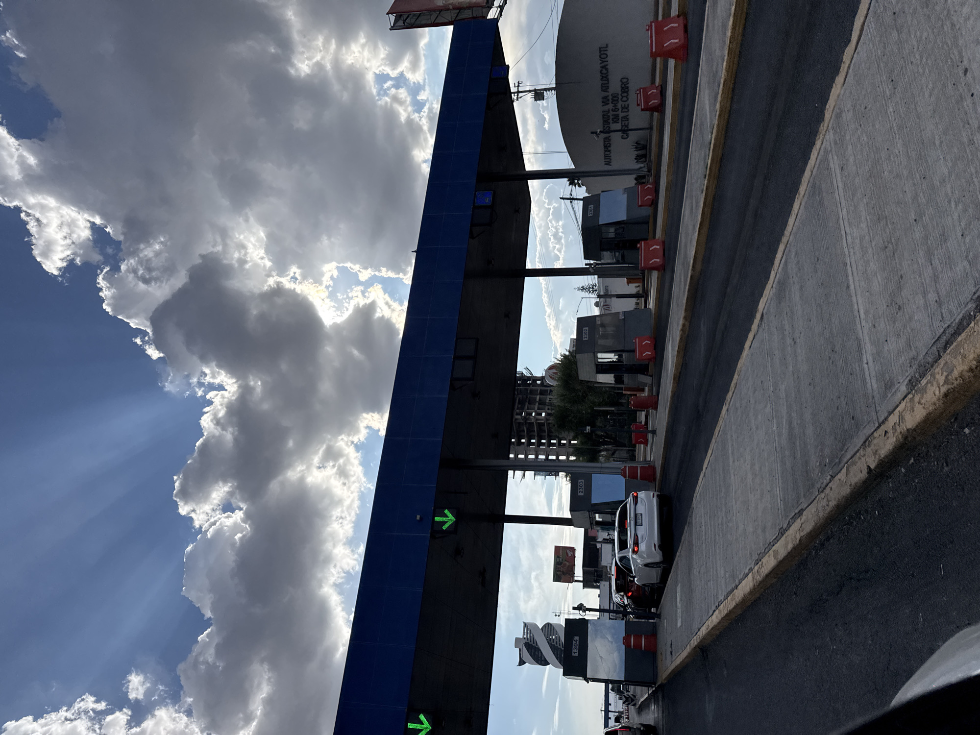 The image depicts a toll booth on the Autopista Estatal Via Atlixcayotl in San Bernardino Tlaxcalancingo, Mexico, at kilometer 6+000. The sky is partly cloudy with sunlight breaking through. The toll booth structure is blue, and there are multiple lanes with red markers. Cars are lined up at the toll booths. A sign indicates "CASETA DE COBRO" (toll booth) and other text is visible including booth numbers. A building and trees are visible in the background. The roadway is gray concrete with a yellow border.