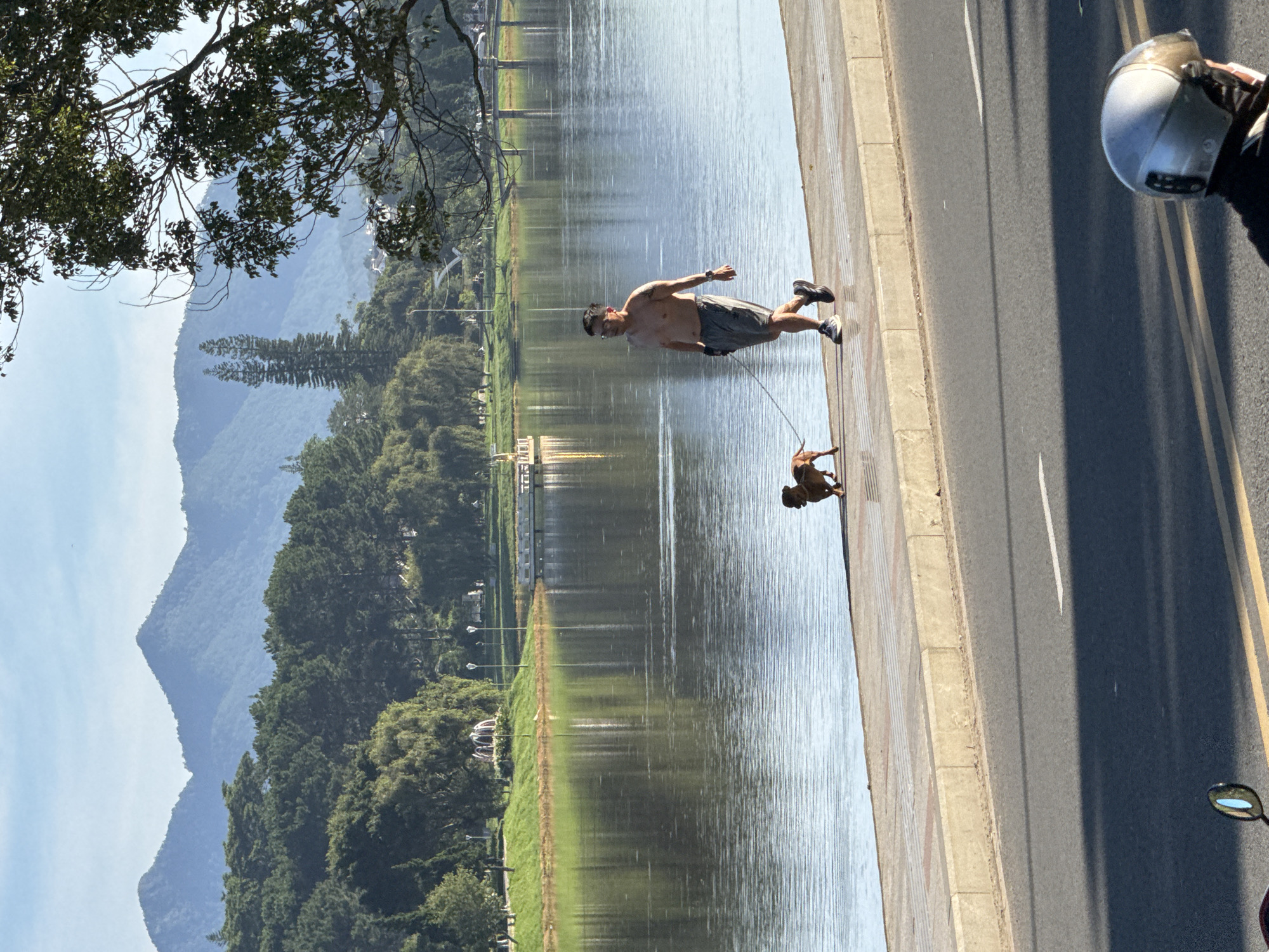 An adult male, shirtless and wearing grey shorts, walks a small brown dog on a leash along the paved walkway next to Xuan Huong Lake in Da Lat, Vietnam. The man is wearing athletic shoes and a watch on his left wrist. The dog is a short-haired breed, possibly a Dachshund. In the foreground, the tarmac road reflects sunlight, with a motorcycle mirror visible in the bottom right corner. Behind the walkway, the lake is calm and reflects the sky and surrounding trees. Across the lake, a green park area features manicured grass, trees, streetlights, and a decorative fountain. In the background, undulating, tree-covered mountains rise against a clear blue sky. The overall scene depicts a peaceful morning activity by the lake.