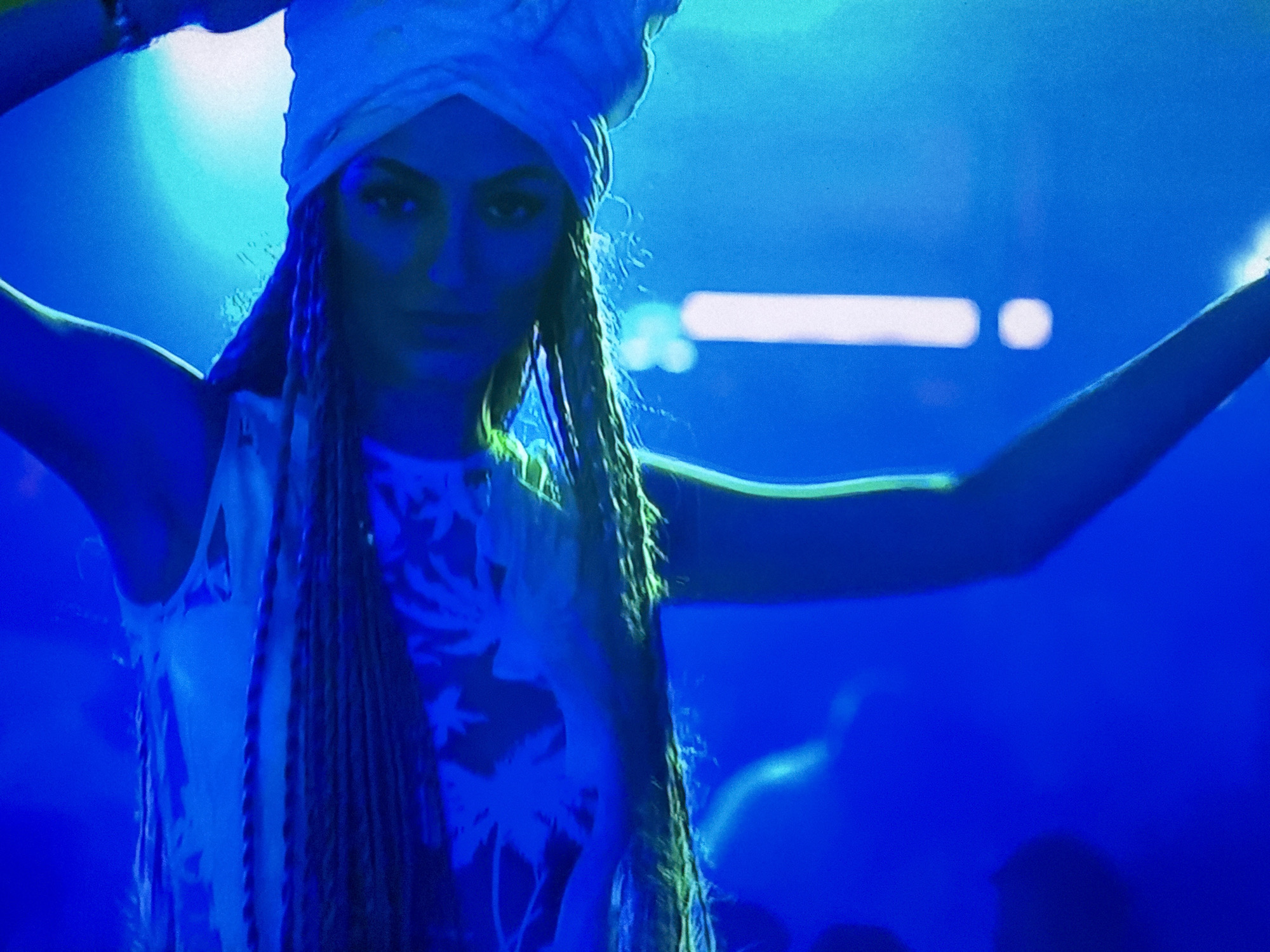 A woman is standing in a dimly lit room in an unknown city. She has her arms outstretched and is wearing a white head wrap, a white tank top with palm tree designs, and long braids. The lighting in the room is predominantly blue, with some green highlights around the woman. There are other people in the background, but they are out of focus.