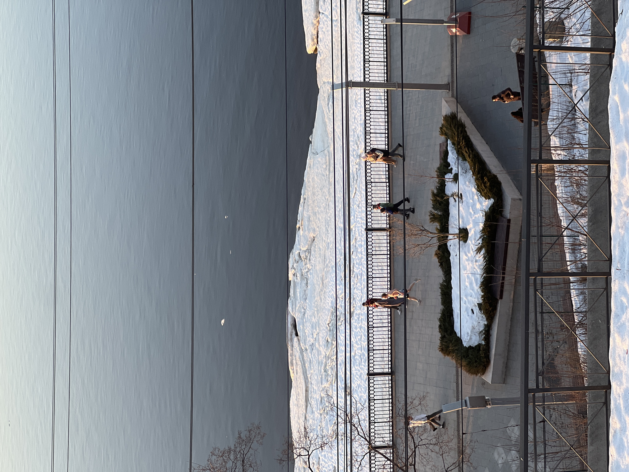 The image depicts a scene in Perm, Russia, specifically the embankment of the Kama River, likely during the winter or early spring season, given the presence of snow and ice. From an elevated, top-down perspective, the view encompasses the river, the embankment promenade, and adjacent urban features.
The left side of the image shows the Kama River, a large expanse of grey water with a small white spot near the center. Along the riverbank is a buildup of snow and ice, indicating cold temperatures. A series of overhead wires spans the scene, running across the river.
The embankment features a paved walkway with a decorative black metal fence running parallel to the river. Several people are walking along the promenade. The composition includes an elevated, rectangular planting bed with greenery and snow covering the surface. Benches are positioned near the edges of the planting bed, as well as along the embankment, and there are lamp posts at certain intervals.
On the right side of the image, the urban environment extends beyond the embankment, with architectural elements such as buildings and fences. Some areas are covered with snow, and bare trees are visible. The angle of the sun suggests that it's early morning or late afternoon.