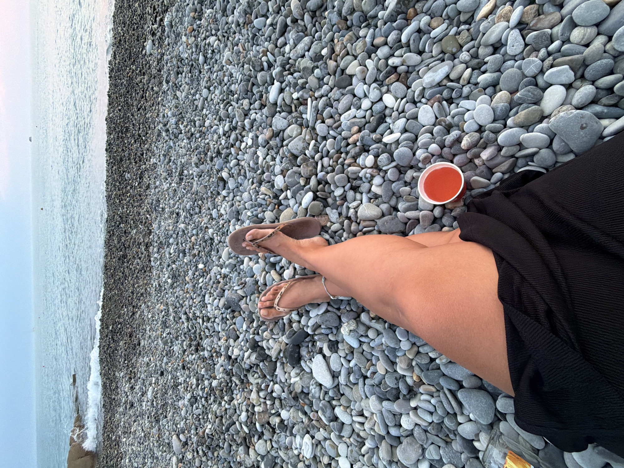 A POV shot shows a person's legs and feet resting on a pebble beach with the sea and horizon in the background. The person is wearing flip-flops and a black skirt. A cup of red liquid sits on the pebbles. The beach is covered in gray and white pebbles. The sea is calm and a few boats are visible in the distance. The sky is overcast. The location is identified as Sochi, Russia.