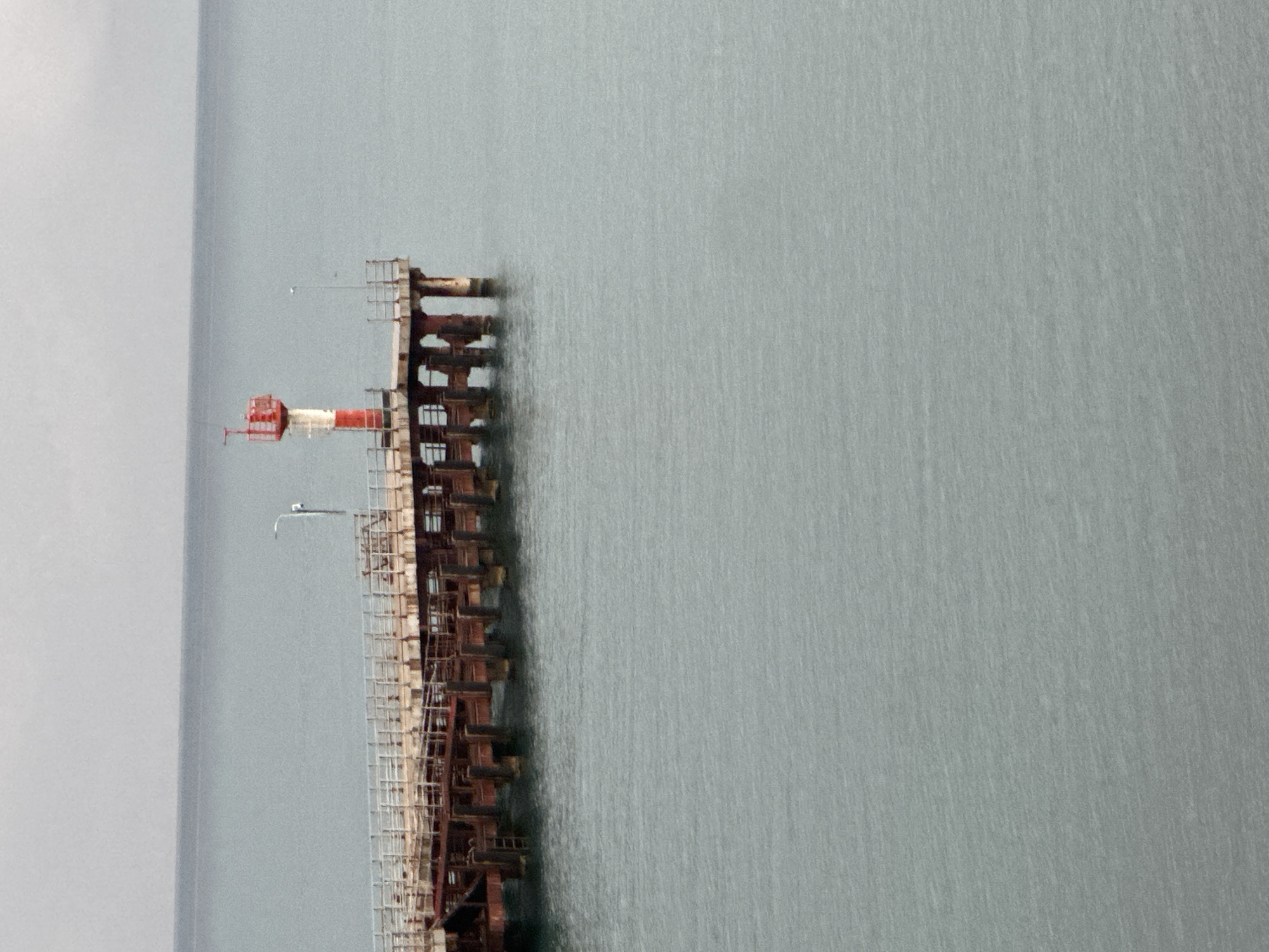 The image shows a pier extending into a calm body of water under an overcast sky. On the pier, a red and white lighthouse stands at the end. The pier itself appears to be constructed of weathered metal and wood, with sturdy support pilings visible in the water. A few lights are mounted on poles along the pier. The water is a muted grey-blue, reflecting the cloudy sky. The scene suggests a functional pier, possibly in a port or harbor, with the lighthouse serving as a navigational aid. This pier is located in Sochi, Russia, specifically on the coast of the Black Sea.
