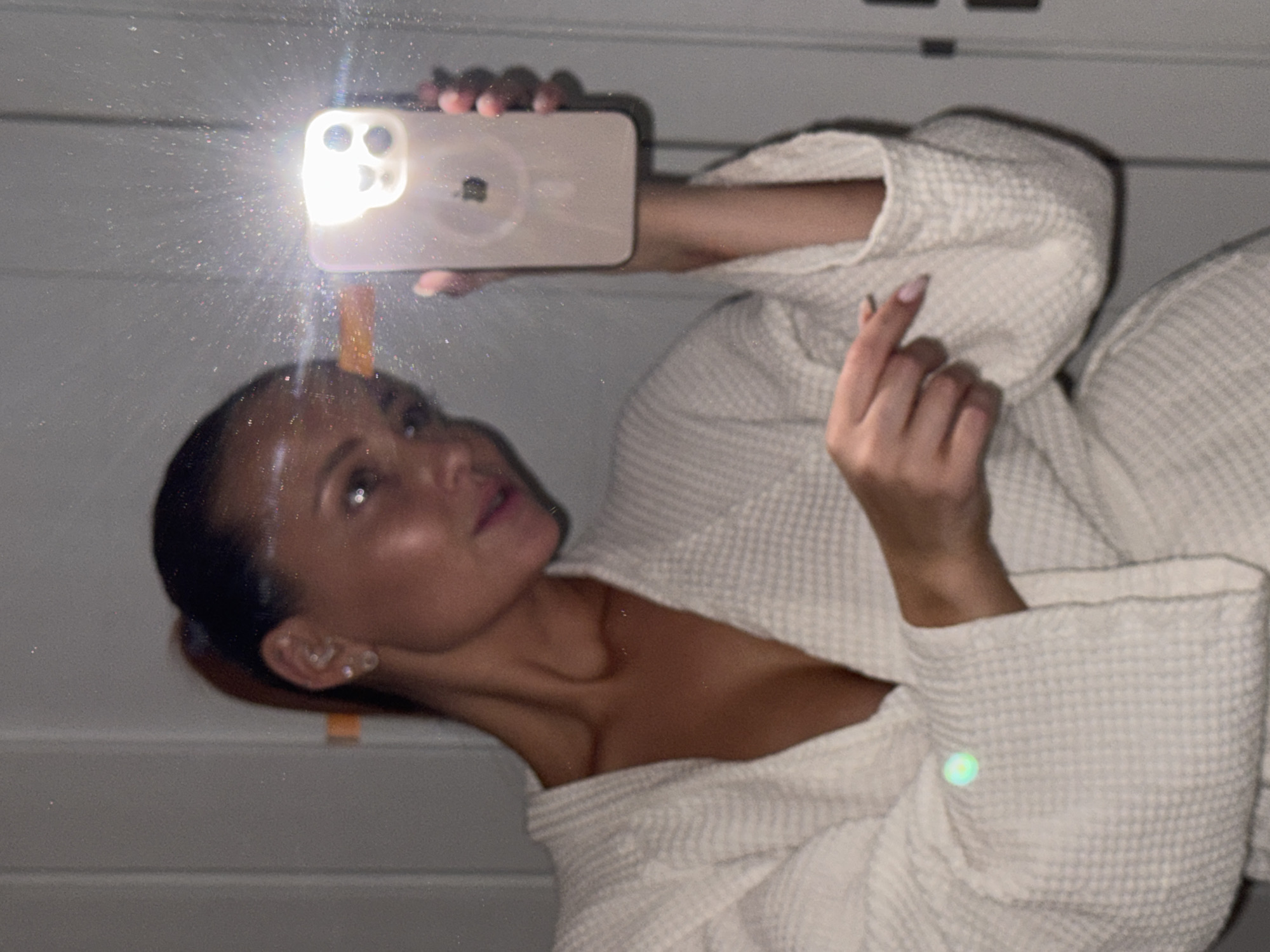 A woman is taking a selfie in a bathroom. She is wearing a white waffle-knit robe. She is holding a phone with its flashlight on, directed toward the mirror. The bathroom has light grey or white walls with horizontal grooved paneling near the bottom. The location in the city of Chaikovsky, Russia, cannot be determined from this image.