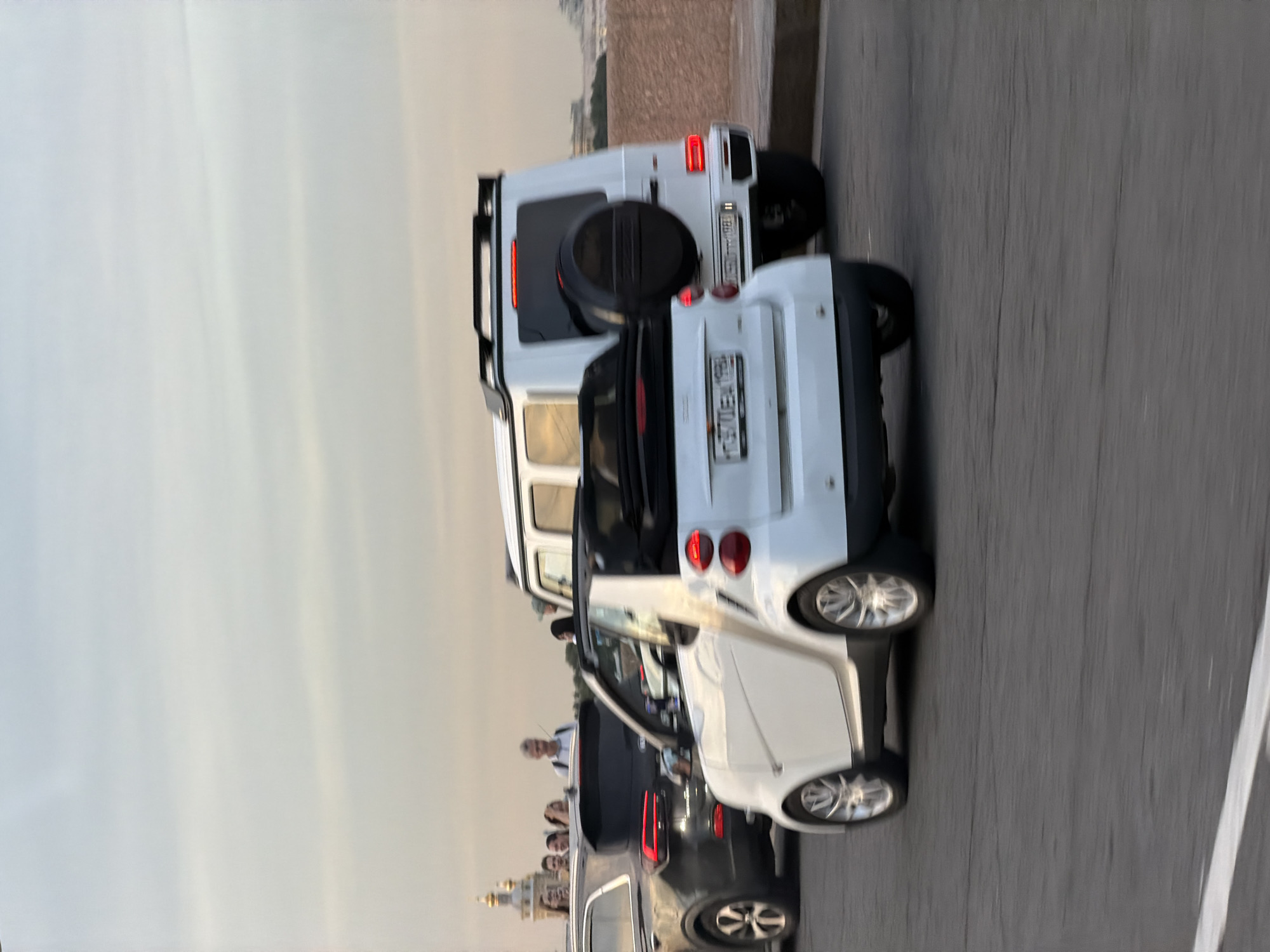 In Saint Petersburg, Russia, on a clear, overcast day, several vehicles are seen on a street. A white Smart convertible is in the foreground, facing left, with its top down. To its right is a white Mercedes-Benz G-Class SUV, facing right. Behind the Smart car, a dark-colored SUV is partially visible. In the background, several people are standing on a raised area, possibly a bridge or embankment, with St. Isaac's Cathedral visible in the distance. The overall scene depicts urban traffic on a bridge with a notable landmark in the background.
