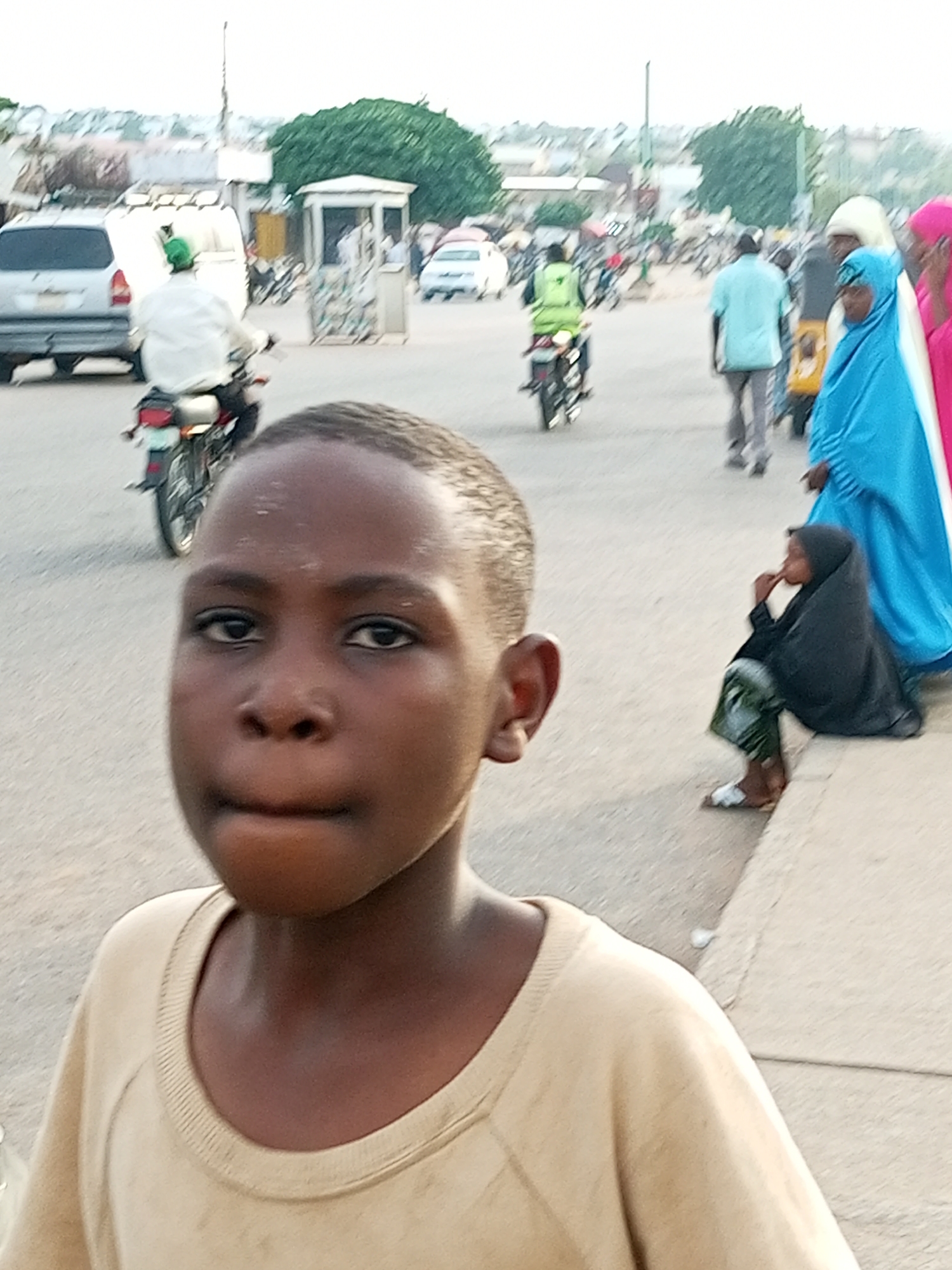The image depicts a street scene in Gombe, Nigeria. In the foreground, a young boy is captured in a close-up shot, looking directly at the camera. He is wearing a light-colored t-shirt. In the background, several people are visible on the street. A person in a bright green vest rides a motorcycle, heading away from the camera. To the right, a woman in a blue hijab and another in a pink hijab are walking. Further to the right, a child in a black hijab sits on the curb, facing left. Various vehicles, including a white van and a silver car, are parked or driving on the road. The environment suggests an urban setting with a paved road and buildings in the distance. The lighting indicates it is daytime.