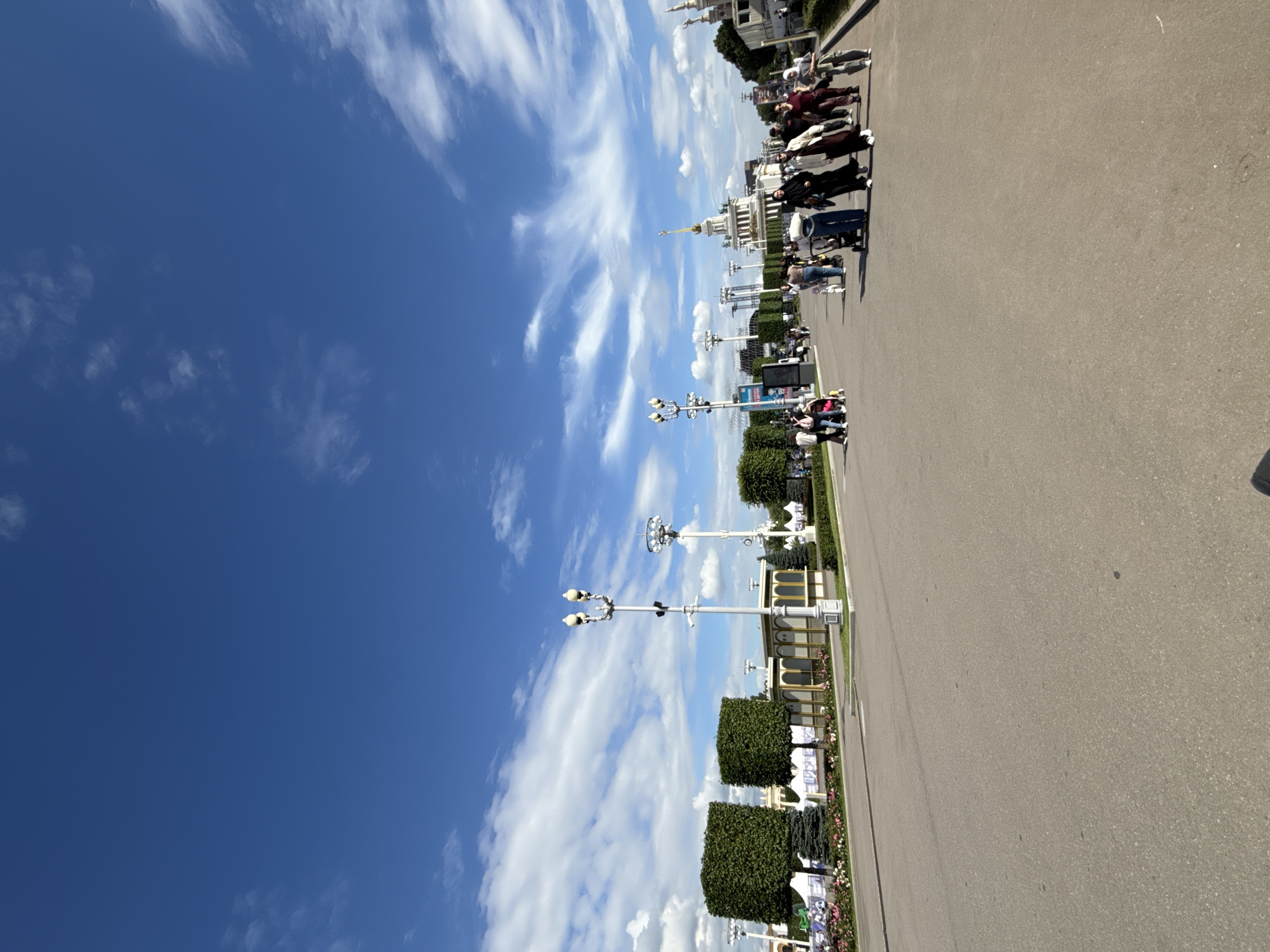 The image depicts a wide, paved pathway in an outdoor urban setting, likely a park or exhibition grounds in Moscow, Russia. The sky is a vibrant blue with scattered white, wispy clouds. On the left side of the pathway, there are manicured, square-shaped topiaries and a row of flowering plants. Several ornate lampposts with multiple lights are spaced along the pathway. In the background, a large white building with a prominent spire and golden dome is visible, a common architectural feature in Moscow, suggesting this might be a location like VDNKh (Exhibition of Achievements of National Economy). A diverse group of people, some dressed in traditional Islamic attire, are walking along the pathway. Several individuals are pushing strollers, indicating a family-friendly atmosphere. The scene is brightly lit by daylight, suggesting a summer or spring day.