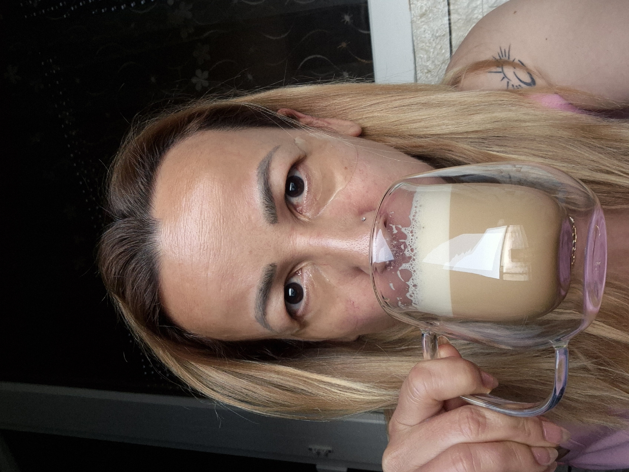 A medium shot shows a person holding a clear double-walled mug of coffee with foam on top to their mouth. The person has blonde hair and is wearing a pink shirt. They are wearing under-eye patches and have a nose ring. The background is out of focus and appears to be indoors with a window or door on the right. The person's left arm is visible as they hold the mug. A tattoo of a sun with a crescent moon is visible on the person's shoulder.