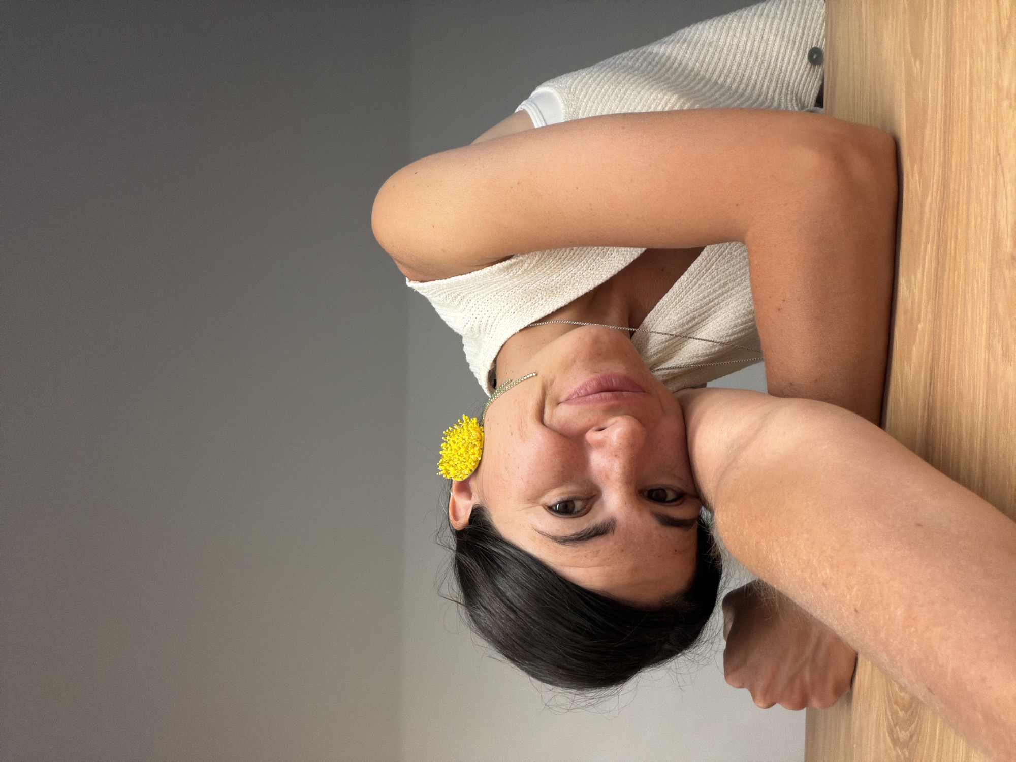 A vertically oriented portrait captures a woman looking directly at the camera with a subtle smile. Her head appears inverted and is positioned on the left side of the frame. She has dark hair and dark eyes. She is wearing a light-colored, possibly off-white or cream, knitted top or sweater. A delicate silver chain necklace adorns her neck, and a prominent, vibrant yellow, spherical earring with a textured, beaded appearance is visible on her left ear. Her arms are bent, and she seems to be resting her head on one arm, which is partially visible on a light-colored wooden surface that occupies the right half of the image. The background behind her head is a plain, light grey wall, suggesting an indoor setting. No text is visible. The overall impression is casual and friendly.
