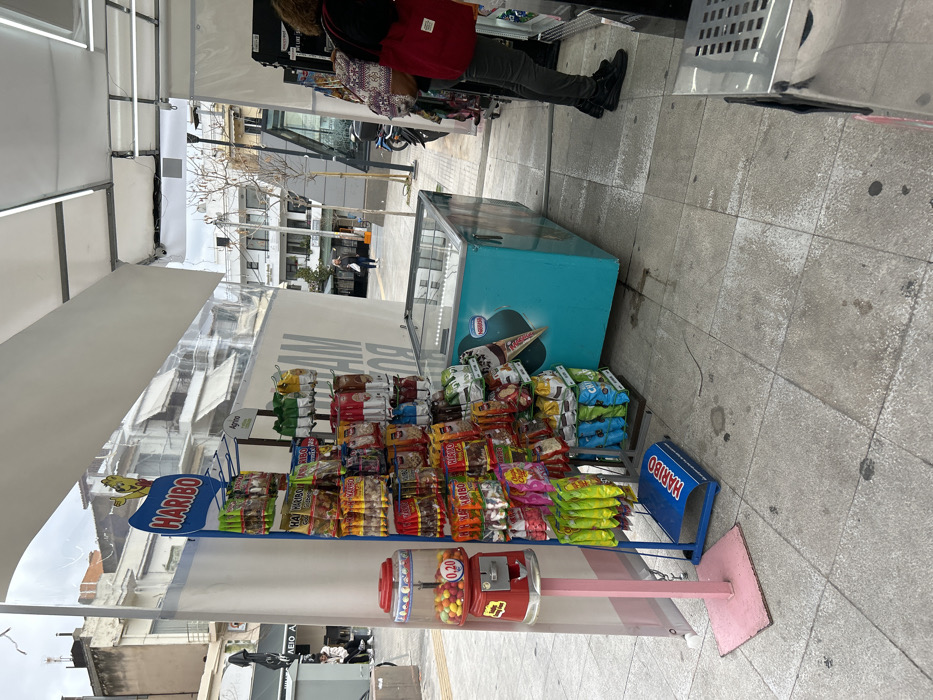 This image captures a vibrant outdoor street vendor stall, likely located in an urban area such as Nikea, Greece. The scene is set on a paved sidewalk, illuminated by bright daylight, suggesting a busy daytime atmosphere.
In the foreground and center, a white canopy tent shelters a well-stocked snack display. Numerous packages of chips, candies, and other treats are neatly arranged on shelves. A prominent blue sign with the "HARIBO" logo and its iconic bear mascot is affixed to the left side of the display, with another smaller blue "HARIBO" sign visible near the bottom right. At the base of the display, a red gumball machine filled with colorful candies proudly shows a price of "0.20," presumably in euros.
To the right of the snack stand, a light blue chest freezer for ice cream is positioned, bearing the "Nestlé" logo with an ice cream cone graphic. Beyond the freezer, a person wearing dark pants, dark boots, and a red backpack stands, seemingly attending to another part of the vendor's setup or browsing.
The ground is a light grey paved surface, showing some signs of use and minor discoloration. In the background, an urban environment unfolds with multi-story buildings, some bare-branched trees, and more paved areas where other people are visible walking in the distance. The overall impression is that of a bustling public space where snacks and treats are readily available from a street vendor. Partial, reversed text that could read "BUY ME" or similar is faintly visible on a banner behind the snack display.