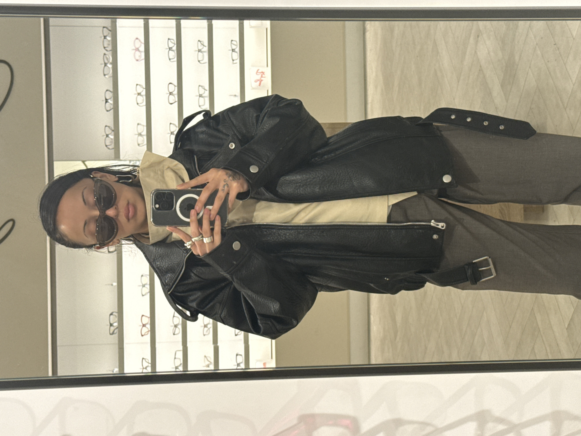 A person is taking a mirror selfie in what appears to be an eyewear store. The person is wearing sunglasses, a light-colored hoodie, and a black leather jacket. Their trousers are gray with a belt detail. The person's left hand, holding the phone, is visible, adorned with rings and tattoos. The background showcases multiple rows of eyeglasses displayed on shelves. The location seems to be an eyewear store, as evidenced by the numerous glasses frames displayed. The scene is brightly lit with artificial lighting. The reflected image suggests the person is standing in front of a large mirror within the store, taking a photo of themselves. It is impossible to verify the exact location of this scene in London from the image.