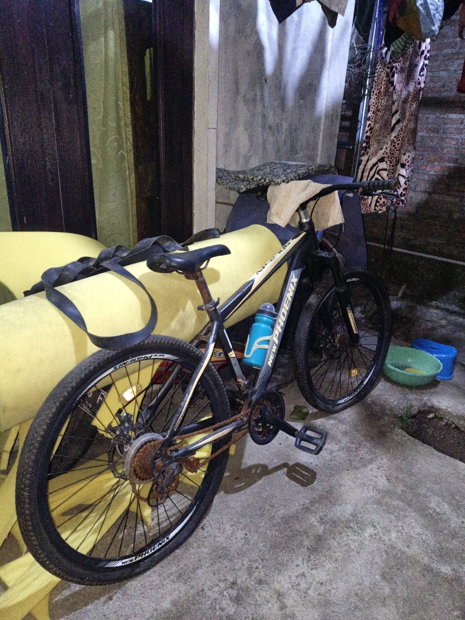A bicycle is leaning against a bright yellow cushioned surface. The bicycle is black with some white and yellow detailing on the frame. A black inner tube is draped across the bicycle and the yellow surface. The bicycle has a blue water bottle attached to the frame and is resting on a concrete ground. In the background, there are dark brown doors and a lighter colored wall, with some clothing hanging. There are also two plastic basins, one green and one blue, on the ground to the right of the bicycle. The scene appears to be outdoors, possibly in a backyard or alleyway, in Cikal, Indonesia. The lighting suggests it might be evening or a dimly lit area.