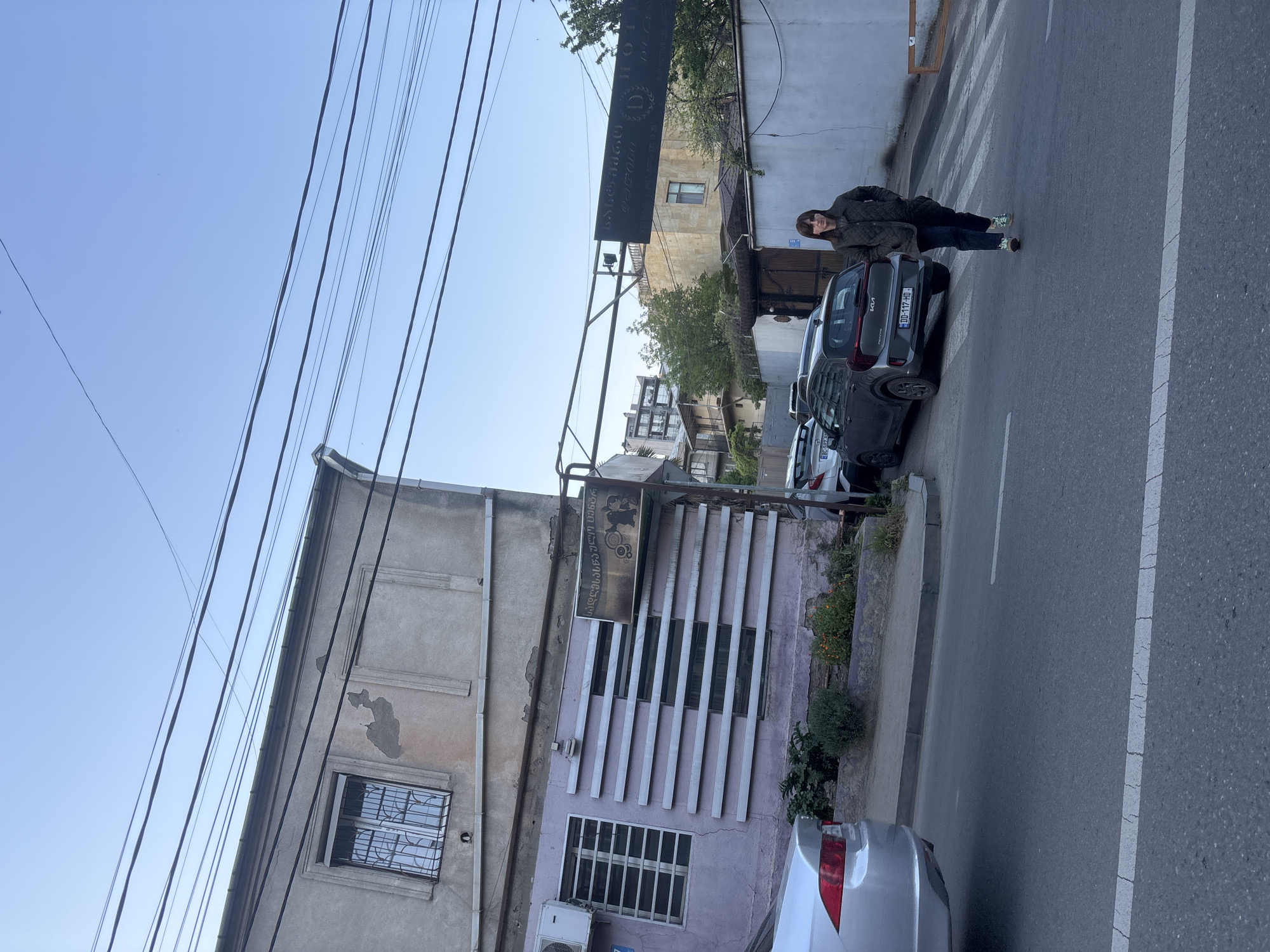 The image captures a daytime street scene in Tbilisi, Georgia, under a clear blue sky. A woman, dressed in a dark jacket and jeans, stands on the sidewalk next to a parked silver Kia Rio. The car's license plate, "DD 117-HD," is clearly visible. The street features white road markings, including a crosswalk.
On the left side of the street, there are several buildings. The leftmost building is an older, two-story structure with weathered, peeling paint and a window secured with metal bars. Adjacent to it is a building with a lighter-colored facade, possibly purple, decorated with prominent vertical slats and multiple windows, some also featuring security bars. An air conditioning unit is visible near the bottom of this building. Overhead, numerous power lines crisscross the sky.
Two signs with Georgian script are notable. A large, black rectangular sign hangs above the street, displaying the Georgian text "ჯონ დიო" (transliterating to "Jon Dio") and a stylized 'D' logo, along with smaller, less legible text. Another sign, mounted on the building with vertical slats, also features Georgian writing; the word "სილამაზის" (beauty) is discernible, strongly suggesting the establishment is a beauty salon. The overall atmosphere is that of a bustling urban environment.
