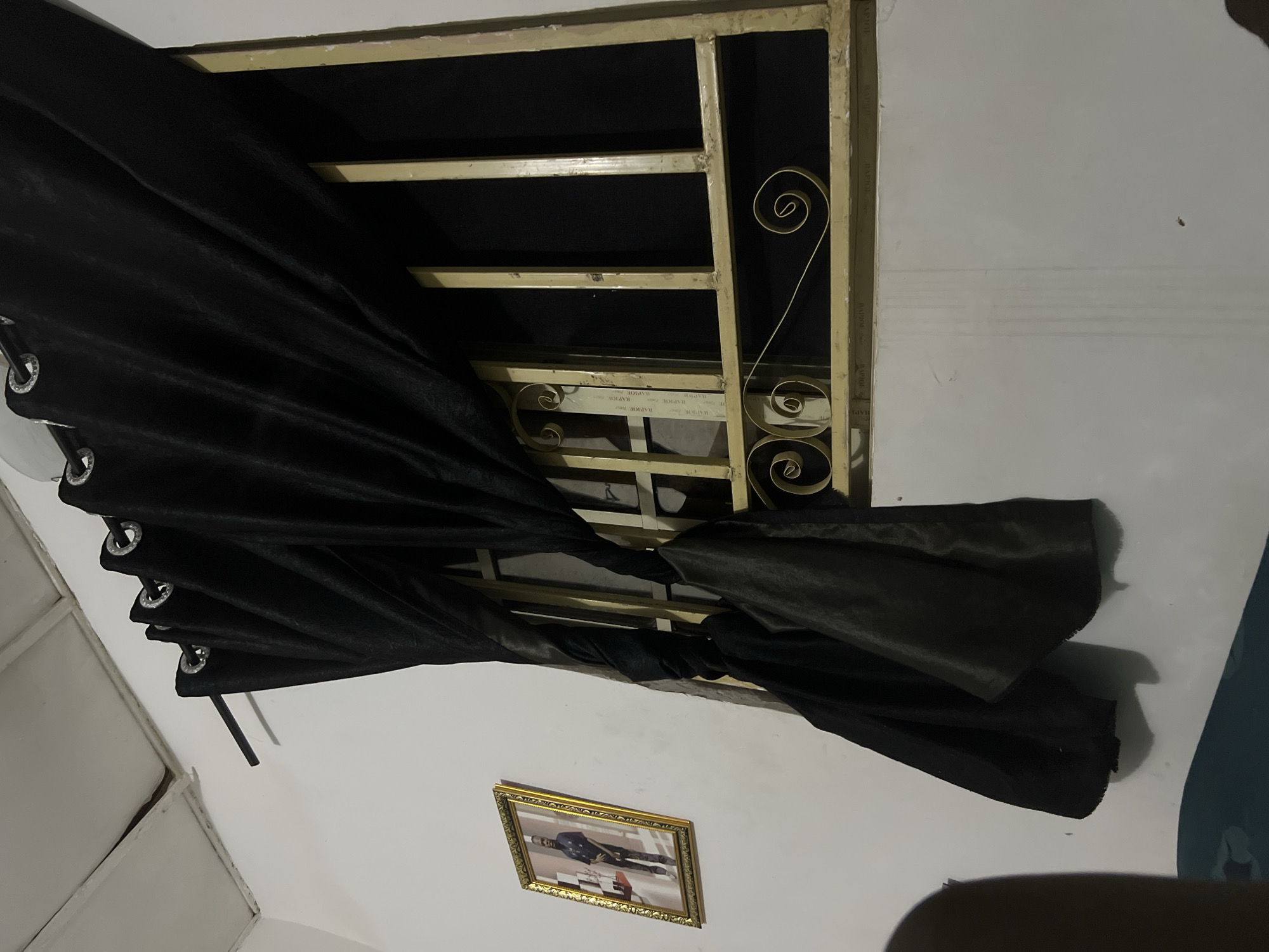 The image depicts an interior scene, likely a room in a residential building in Howayi, Nigeria. A black curtain is drawn partially across a window, which is fitted with a metal security grate. The grate is gold-colored and features decorative scrollwork. To the left of the window, a framed photograph hangs on the white wall. The photograph shows a man in uniform, standing. The ceiling is visible at the top of the image, with visible panels and some signs of wear. The overall lighting suggests it is indoors, possibly at night or in a dimly lit room. There is no visible activity or interaction occurring in the scene.