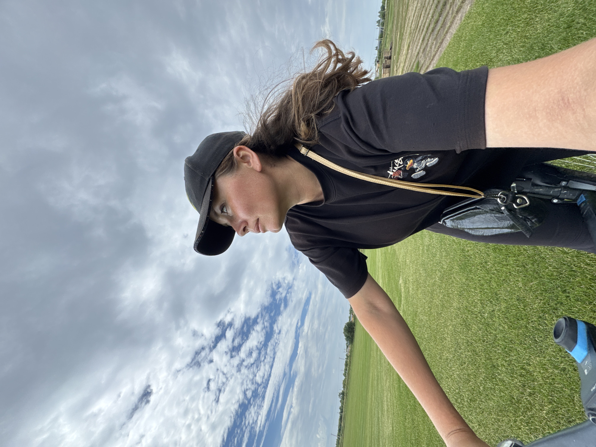 A young woman wearing a black t-shirt and a black baseball cap is on a bicycle, looking up and to the right. She has long, brown, wavy hair that is blowing in the wind. She is wearing a small black purse with a gold strap over her shoulder. The background is a bright green grassy field with rows of crops in the distance. The sky is cloudy with patches of blue sky visible. The image appears to be taken outdoors in a rural area. The specific location of Kempelevo, Russia, is not evident from the visual information in the image.