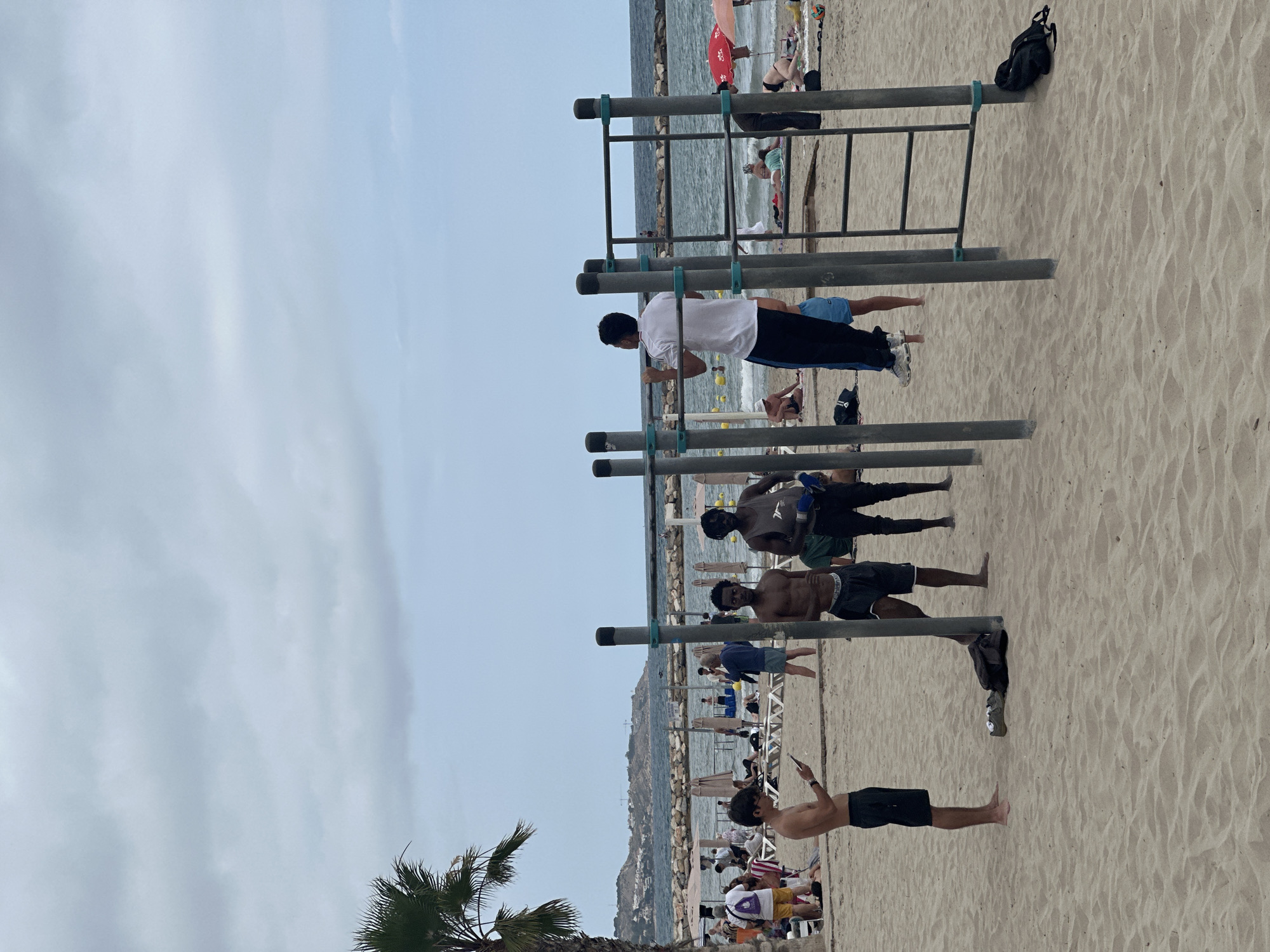 The image depicts a sandy beach with a cloudy sky overhead. In the foreground, a group of young men are engaged in various activities around a calisthenics structure. One man, wearing black shorts, is holding a smartphone while standing on the sand. Two other men, one bare-chested and wearing dark shorts, and another in a grey tank top and dark shorts, are standing near the structure. A fourth man, wearing a white polo shirt and black pants with blue accents, is performing a pull-up on the structure. Further back on the beach, numerous people are relaxing on lounge chairs, under umbrellas, and near the water. The ocean is visible in the background, with a rocky breakwater extending into the water. Palm trees are also present on the left side of the image. The overall scene suggests a casual day at the beach with opportunities for exercise and recreation.