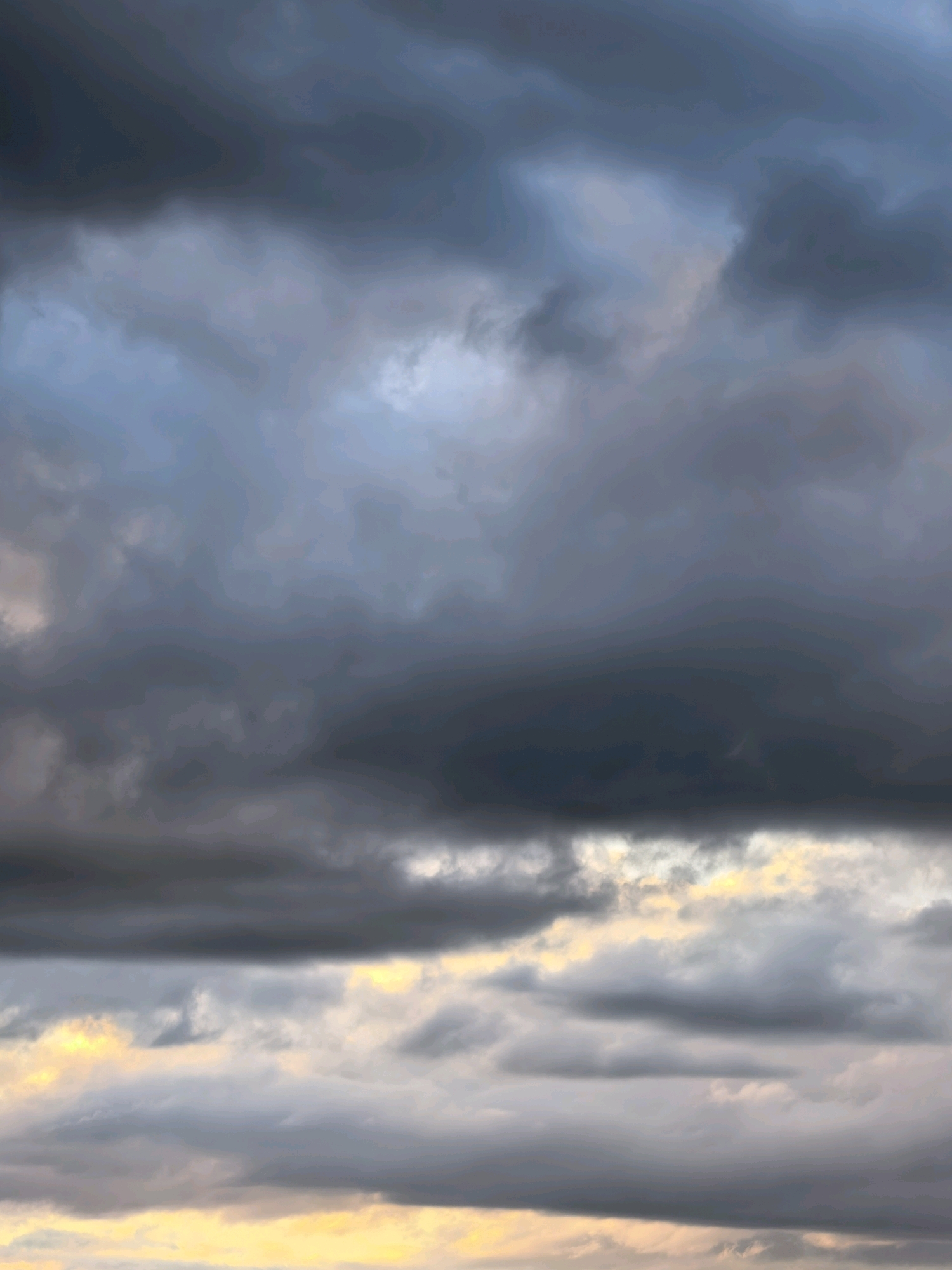 The image displays a dramatic sky filled with heavy, dark, and gray clouds. The clouds are layered, with some appearing denser and more ominous than others. Patches of lighter sky, possibly indicating breaks in the cloud cover or the approaching sunset, are visible towards the bottom of the frame, casting a faint yellow and orange glow. The overall impression is one of an impending storm or a turbulent weather front. There are no visible structures, people, or any discernible elements of a city.