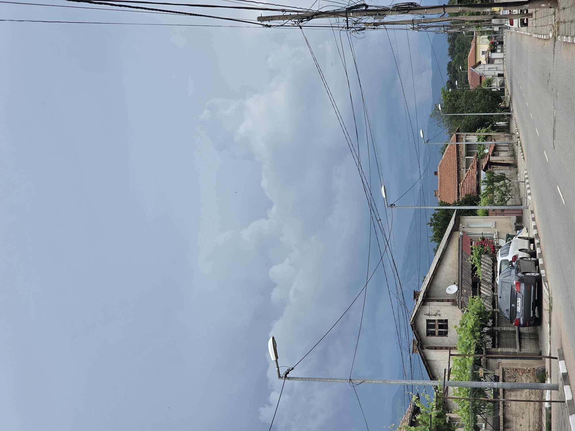 The image depicts a street scene in Aleksandrovo, Bulgaria. A gray asphalt road runs through the frame, flanked by sidewalks with distinctive black and white curbs. Several houses with tiled roofs line the street, some appearing older and weathered. Green trees and vegetation are visible around the properties. Utility poles with numerous overhead wires are a prominent feature along the roadside. A few cars are parked on the side of the road, including a dark gray sedan and a white SUV. Streetlights are present along the street. The sky is overcast with dramatic, heavy clouds, suggesting impending or recent rain. In the background, distant hills or mountains are visible through the haze. No people are visible in the frame.