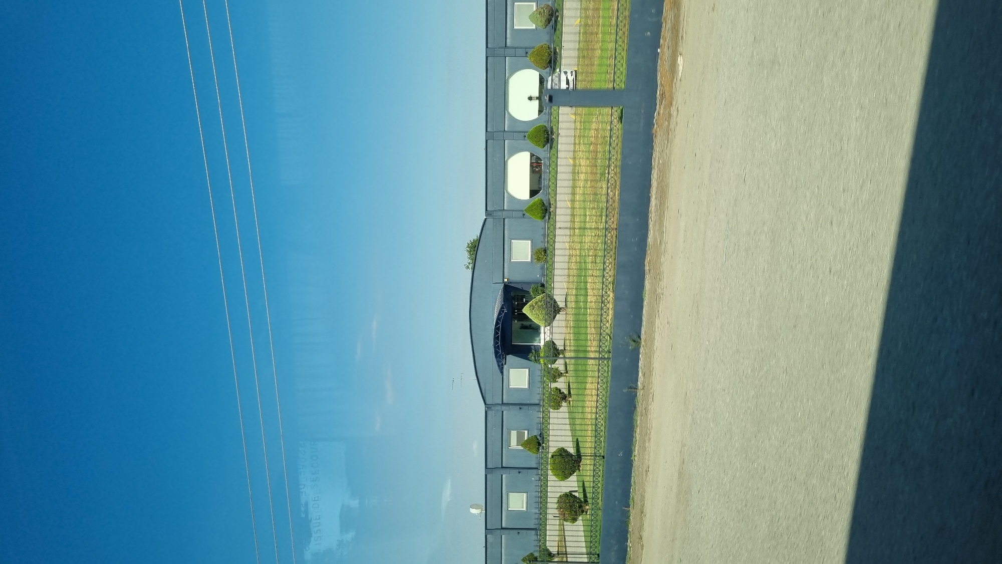 A long, low, grey building with a curved central section is visible under a clear blue sky. The building has multiple rectangular windows and two large circular windows on the right side. A dark blue awning extends over the entrance. In front of the building is a manicured lawn with several cone-shaped evergreen bushes. A dark grey metal fence encloses the property, and behind the fence is a grey asphalt surface. Power lines are visible in the upper portion of the image.