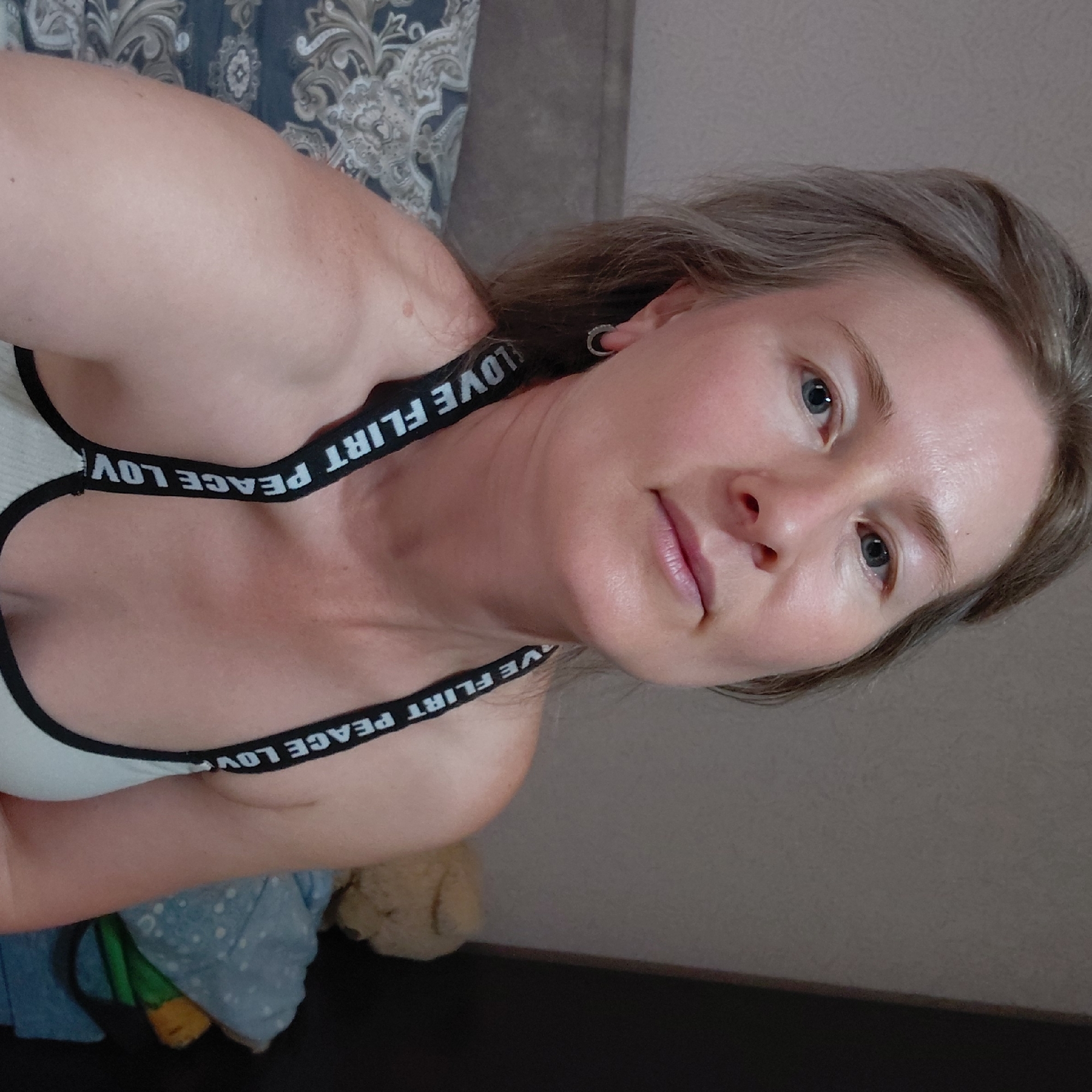 A medium shot shows a woman with light-colored hair, blue eyes, and fair skin, looking directly at the camera. She is wearing a white tank top with black straps that have white text printed on them, spelling out "LOVE FLIRT PEACE LOVE". She has a small hoop earring in her left ear. The background is softly blurred and appears to be a bedroom, with a headboard of a bed visible on the left and a plain wall behind her. A teddy bear and some blue fabric are visible in the background on the right. The lighting is soft and natural. The image was taken in Novosibirsk, Russia.