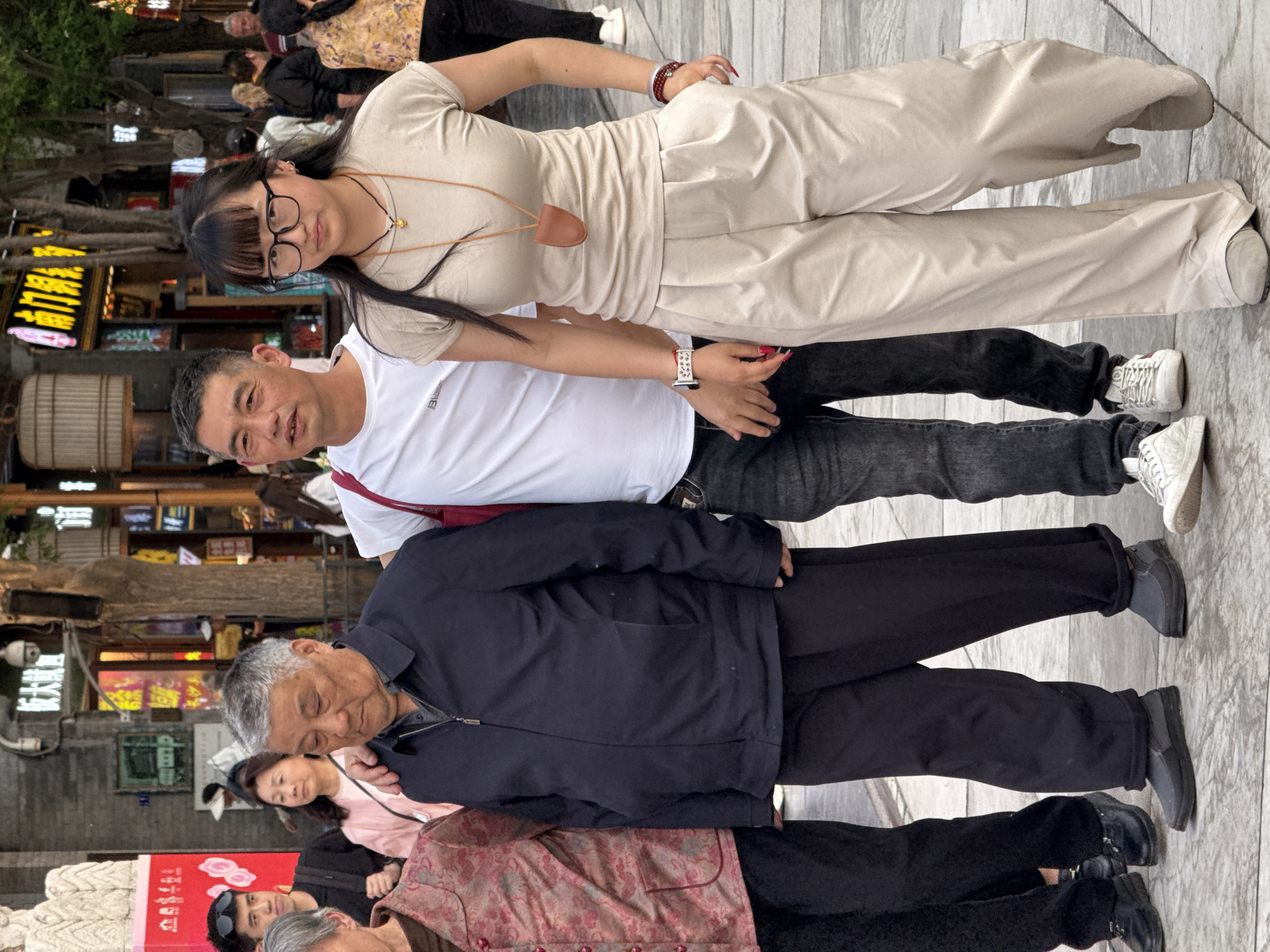 The image depicts a group of people standing in a public space in Dongcheng District, China. The flooring consists of light gray, linear tiles. A young woman with dark hair and glasses, wearing a beige top and pants, stands next to a man in a white t-shirt and black jeans. An older man in a navy jacket and black pants is adjacent to them. Other people, including an older woman in a floral top and dark pants, are partially visible in the background. The surroundings include buildings and trees, with various signs and shopfronts visible.