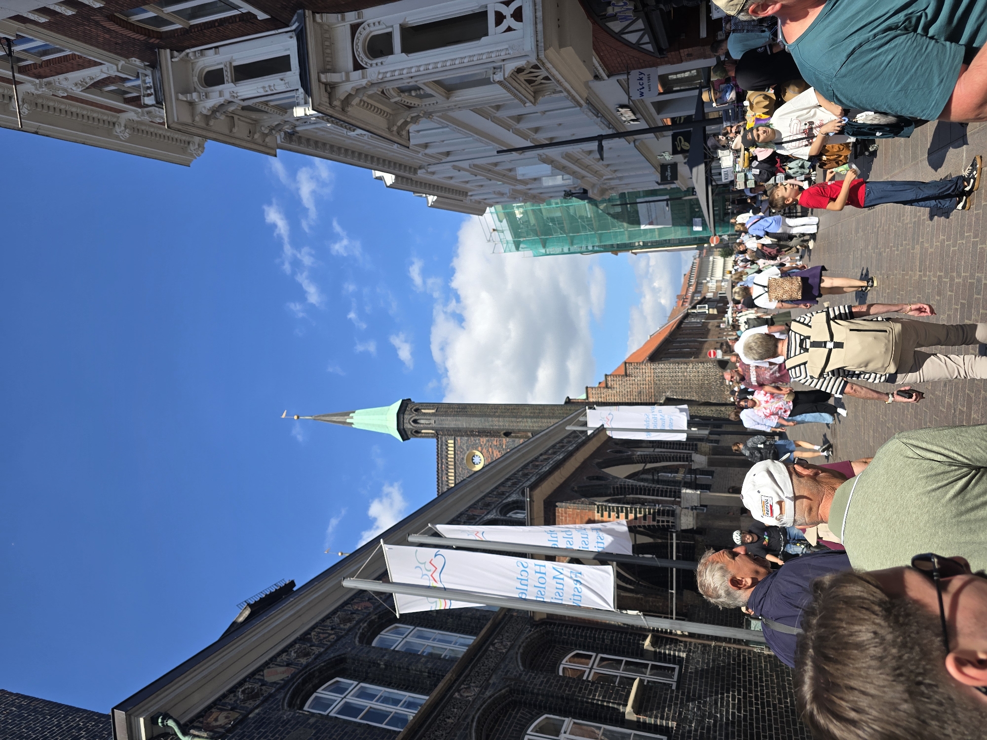 This is a street-level, eye-level, wide-angle shot of a bustling pedestrian street in Lübeck, Germany, specifically likely the Breite Straße, a well-known shopping area. The scene is captured on a bright, sunny day with a clear blue sky dotted with scattered white clouds.
In the foreground, several people are walking and interacting. On the left, a man with graying hair wearing a dark blue jacket is speaking, with the back of another man's head, wearing sunglasses and a dark shirt, visible in front of him. To the right of them, a man wearing a white baseball cap and a green shirt is also facing away from the camera. Further back, a diverse crowd of people walk along the cobblestone street, some browsing market stalls set up along the sides.
The architecture on both sides of the street is historic. The left side features a dark brick building with arched windows, identified as likely part of St. Mary's Church (Marienkirche) in Lübeck, indicated by the tall, green-spired church tower visible in the mid-ground. Several tall, white banners with colorful abstract designs and text are hanging from poles in front of this building. The right side of the street is lined with ornate, white-facaded buildings with intricate detailing and arched windows. One of these buildings displays a sign for "wicky seit 1985". Construction netting covers a building further down the street on the right.
The overall impression is of a vibrant urban environment, a popular public space filled with activity and historical charm.