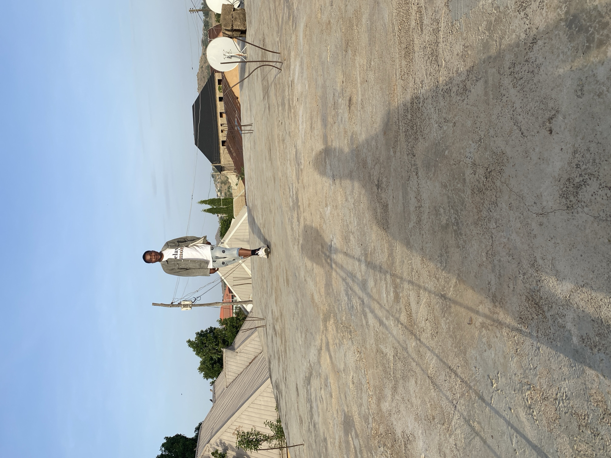 A medium shot captures a dark-skinned male standing on a concrete rooftop in Bukuru, Nigeria, under clear, blue skies. He is facing the viewer and is wearing a light-colored t-shirt with black writing, a jacket, and patterned denim shorts. A long shadow is cast by the man extending out from him across the concrete rooftop. The setting includes satellite dishes, buildings, trees, and telephone poles.