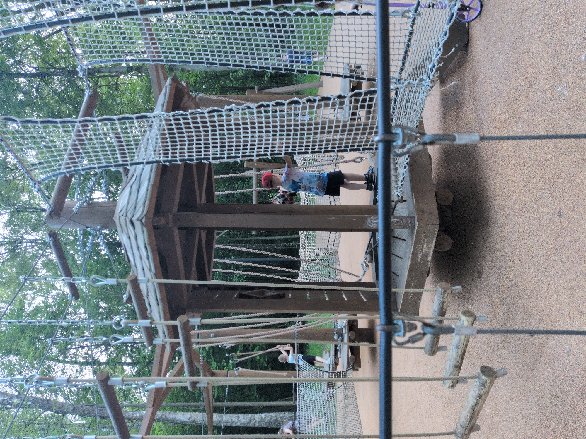 The image depicts an outdoor ropes course and adventure playground structure, likely located in a park or recreational area in Pelikhovo, Russia. The structure is primarily made of wood and rope, with multiple levels, bridges, and climbing elements. In the foreground, a black horizontal bar with a metal loop and cable hangs down, partially obscuring the view. Beyond this, a series of wooden logs suspended by ropes form a challenging walkway.
The central element of the scene is a wooden gazebo-like structure with a hexagonal roof. Inside this structure, a young boy wearing a red cap, a t-shirt with a cartoon character print, and dark shorts stands with his hands on a rope net. He appears to be navigating the ropes course. Other individuals are visible in the background, suggesting a public recreational space. To the left, a woman and child are walking on a net-covered platform. Further back, another child in a blue shirt is visible on a similar platform.
The ground surface of the playground is covered in a soft, sandy material. The surrounding environment is a dense forest with tall trees, indicating the location is within a natural setting. The overall impression is one of an active and engaging outdoor play environment designed for children.
