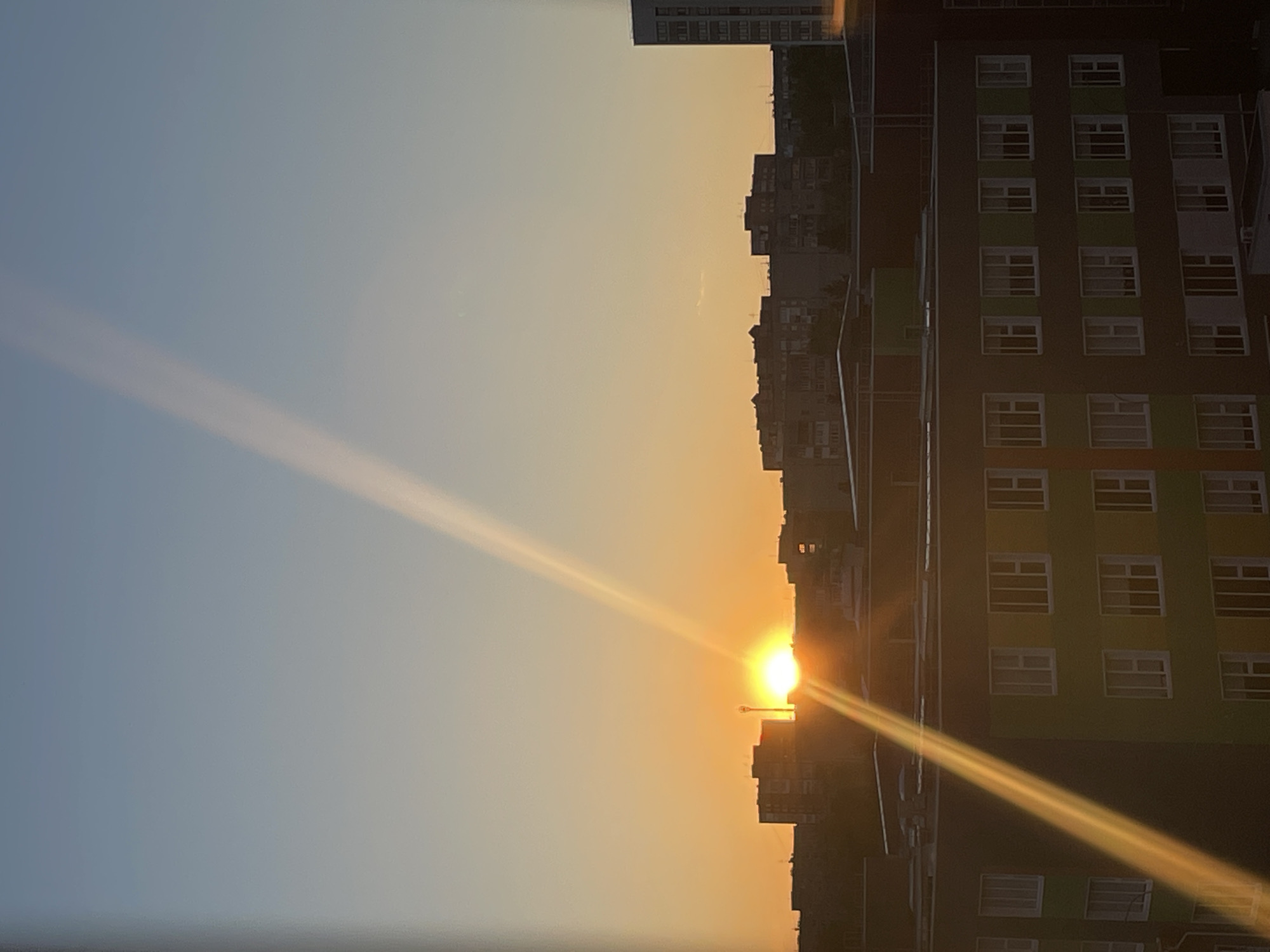 The image shows the sun setting over the city of Izhevsk, Russia. The sun is positioned low on the horizon, casting a bright glare and long rays of light across the sky and buildings. The sky is a gradient of light blue to orange as it nears the horizon. Silhouetted against the sun are several multi-story buildings of various sizes. A modern residential building with many windows is prominent on the right side of the image.
